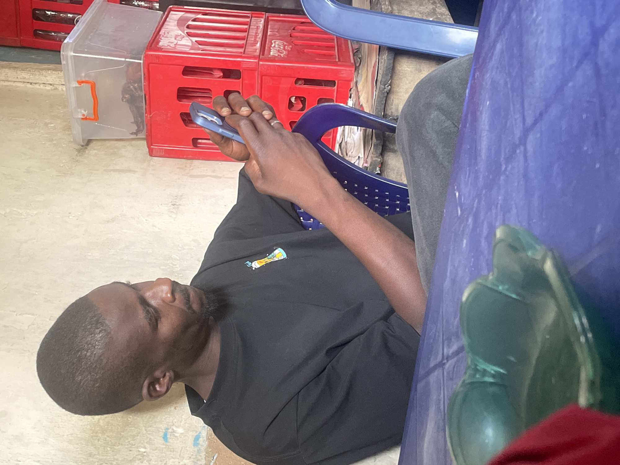 A person is sitting on a purple chair and using a blue mobile phone. The person is wearing a black t-shirt. Adjacent to the person are red crates, one with the Coca-Cola logo. The scene appears to be in Jos, Nigeria.