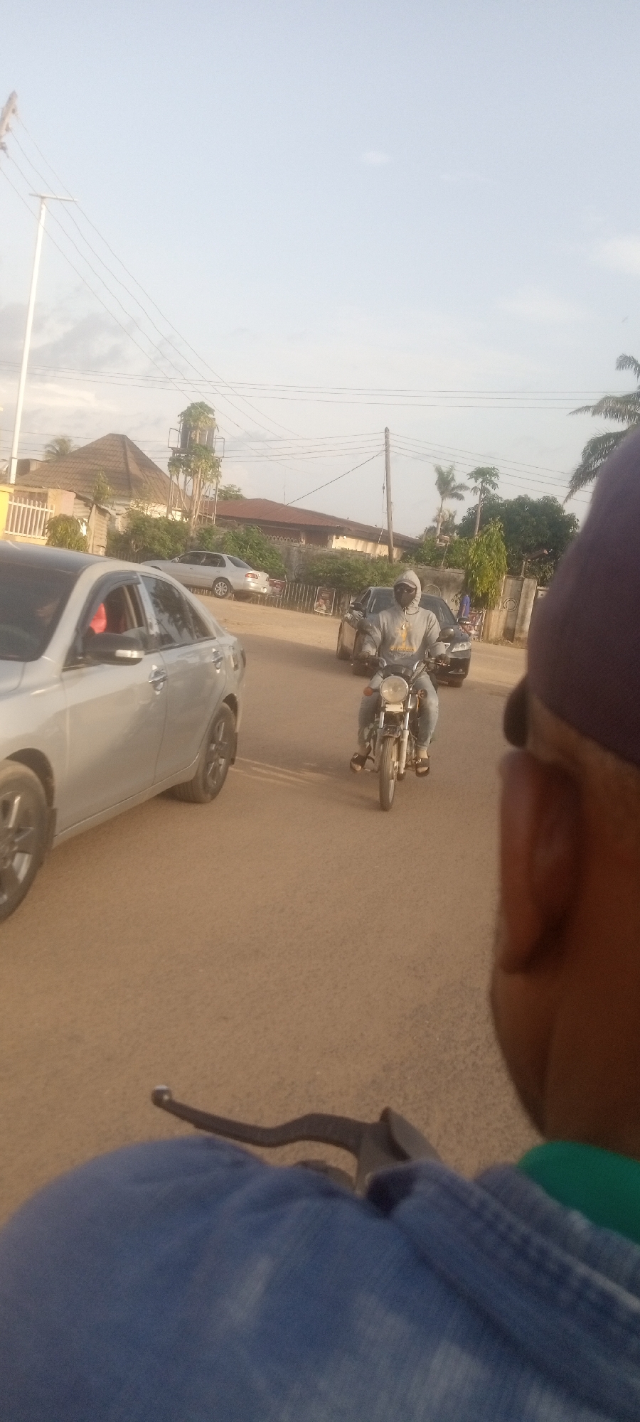 The image depicts a street scene in Narayi, Nigeria. In the foreground, a person wearing blue and a purple baseball cap is riding on a motorcycle. Another motorcycle is approaching in front. A silver car is to the left of the frame. In the background, a couple of houses and other cars are visible. Utility poles and overhead wires are also present. The street appears to be unpaved.
