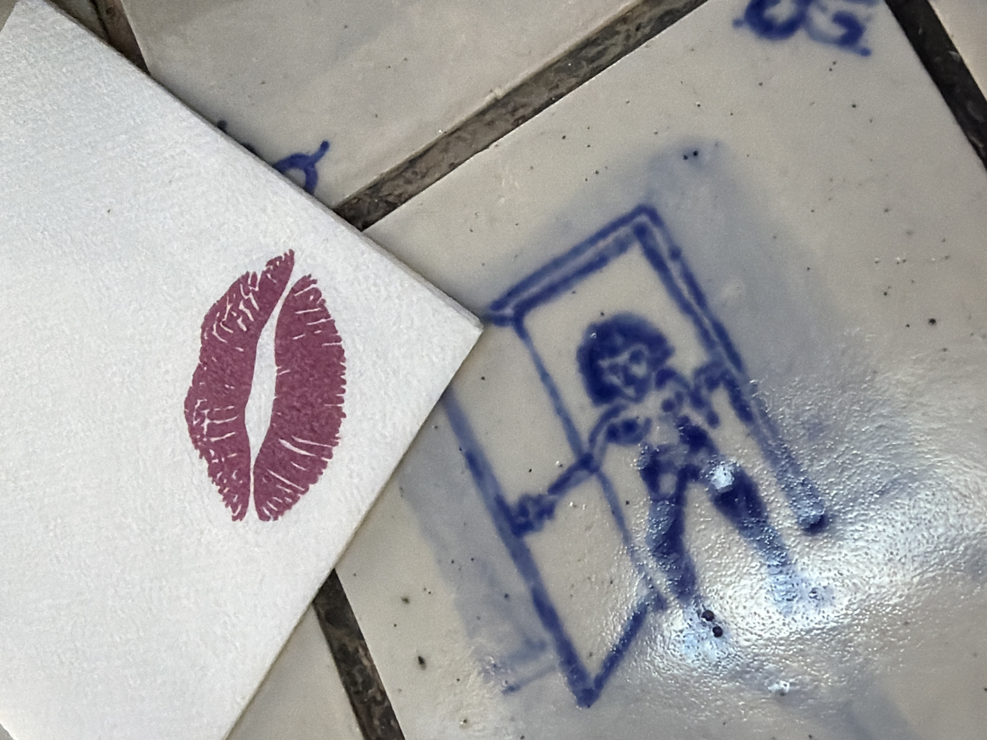 The image captures a close-up, high-angle perspective of a white paper napkin featuring a vivid, magenta-red lipstick kiss print, partially overlaying a tiled surface. The napkin, positioned towards the left of the frame, displays a subtle texture. The prominent kiss mark clearly shows the distinct outline of upper and lower lips with fine detail.
Beneath and to the right of the napkin, light-colored ceramic tiles are visible, separated by darker grout lines. These tiles are adorned with hand-drawn or stenciled blue designs. The most distinct design, located on the lower-right tile, depicts a stylized human figure. This figure, rendered in blue lines, stands within a rectangular frame or doorway, appearing to push against or hold the sides. The tile surface exhibits a slight sheen and scattered small dark specks. Other partial blue markings are visible on adjacent tiles in the upper right.
The scene is static, presenting an arrangement of objects rather than an ongoing activity. The lipstick print suggests a casual, affectionate, or playful moment. The tiles, with their folk-art like blue designs, lend a rustic or artisanal character to the setting. While the image itself does not provide specific location cues, the context provided indicates this scene is in Cape Town, South Africa. No discernible text is present, only graphical elements.