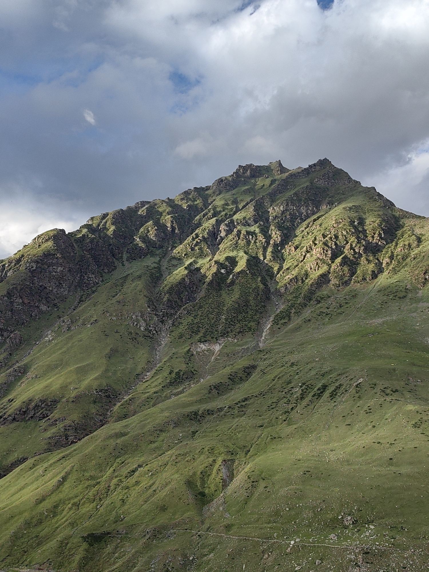 The image shows a steep, green, mountainous slope under a cloudy sky. The vegetation on the slope is predominantly grass, with some rocky outcrops and patches of darker shrubbery. The terrain is uneven, with visible erosion channels and gullies running down the mountainside. The lighting suggests it is either early morning or late afternoon, with shadows cast across the landscape, highlighting the contours of the mountain. In the upper portion of the image, the sky is a mix of dark and light gray clouds, with a few patches of blue visible. The specific location is Gramphu, India. There are no people or man-made objects clearly visible in the frame. The scene depicts a natural, rugged mountain environment.