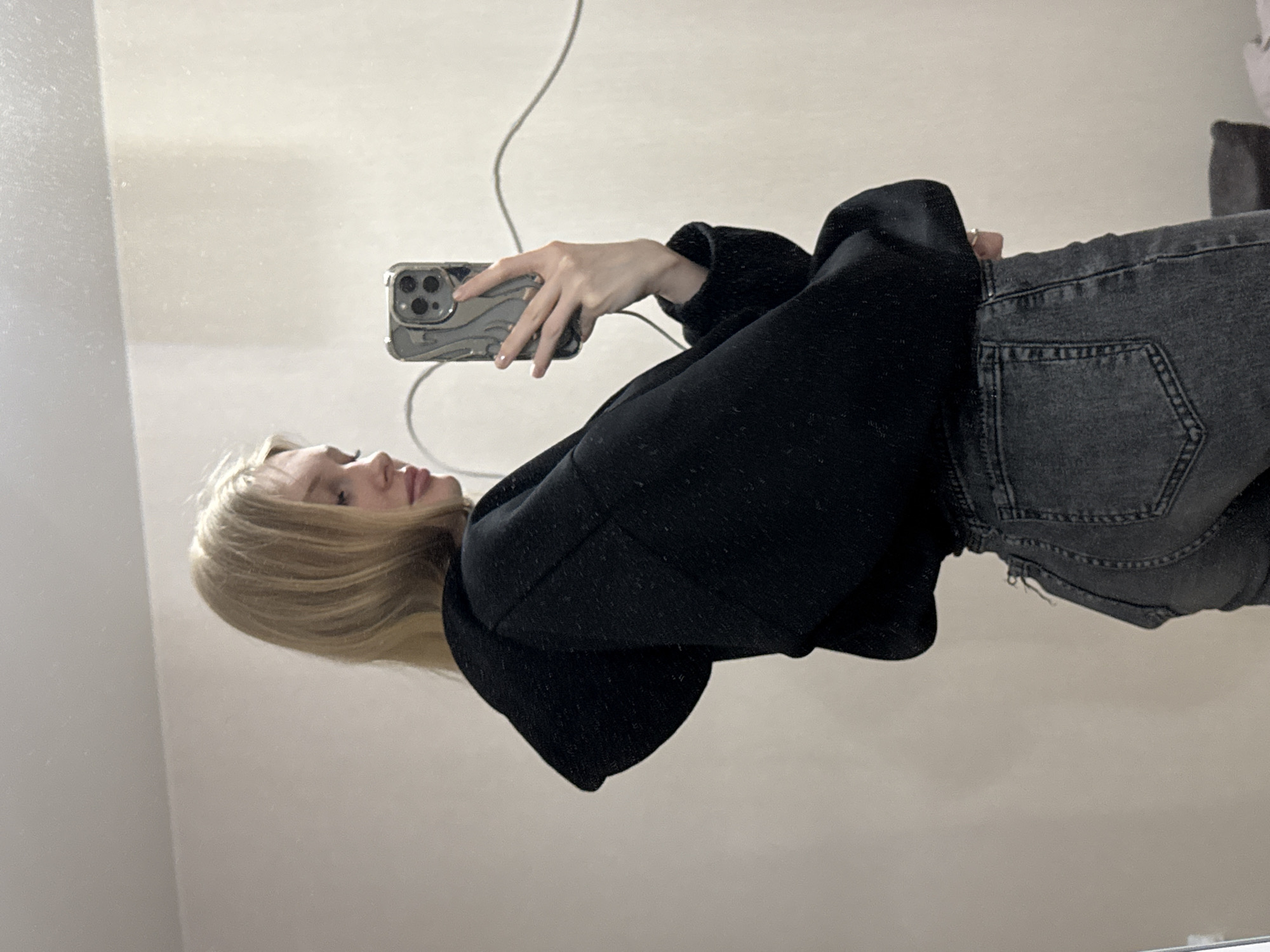 A fair-skinned young woman with long blonde hair is taking a selfie in a mirror. She is wearing a black hooded sweatshirt and grey jeans. She holds a silver phone with a multi-camera lens system in her right hand, angled to capture her reflection. The background shows a plain, light-colored wall. The setting appears to be indoors, likely a private residence. The location cannot be determined as being in Perm, Russia.
