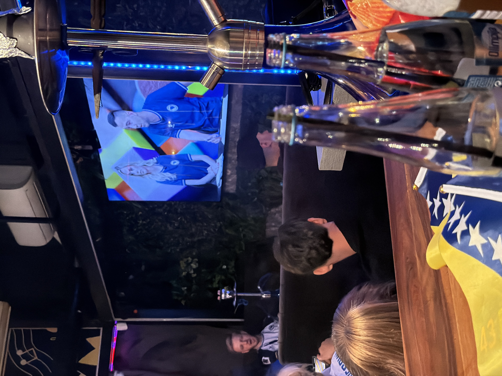 The image depicts an indoor scene, likely a lounge or cafe, with a shisha pipe prominently featured in the foreground. Two glass bottles with blue caps are also visible on a wooden surface. In the background, a large screen displays two individuals, a woman and a man, wearing blue sportswear. Another man with blond hair and a white shirt is seated to the left of the screen, with a shisha pipe behind him. The environment appears dimly lit, with some artificial lighting creating reflections on the glass bottles and the shisha pipe. A flag, partially visible, with blue and yellow colors and white stars, is placed on the wooden surface, suggesting a possible connection to Bosnia and Herzegovina. The scene does not provide specific clues to pinpoint the exact location within the city of Travnik, Bosnia and Herzegovina, beyond the presence of the flag which may allude to national affiliation.