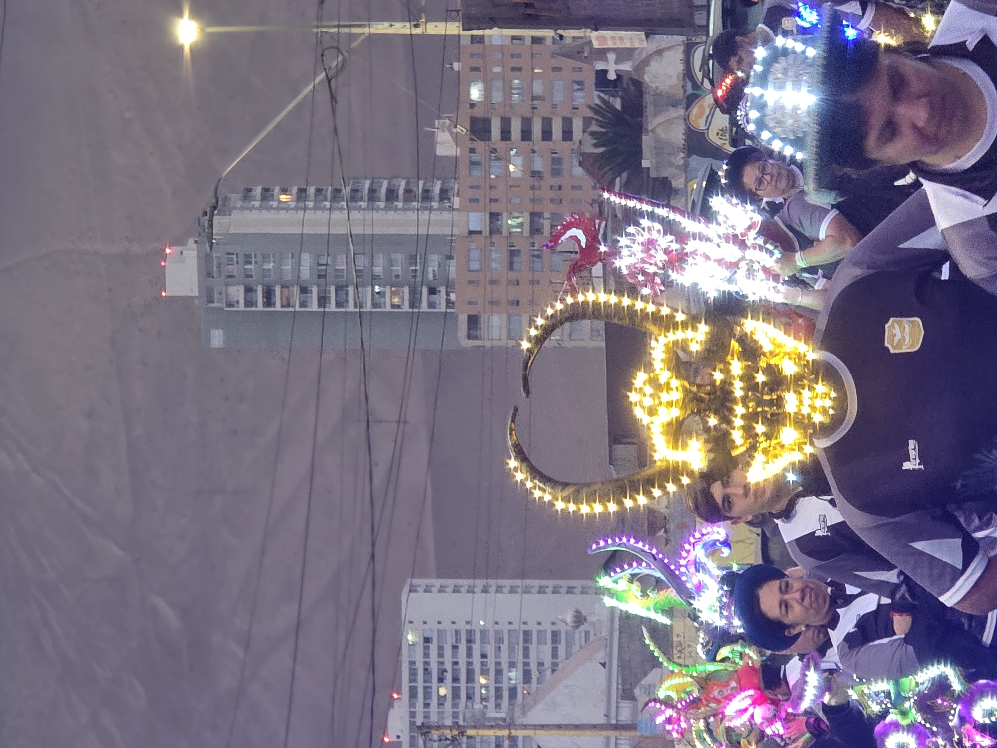 In the city of Iquique, Chile, a nighttime parade is underway, illuminated by vibrant lights. The foreground is dominated by a person wearing a large, horn-shaped mask adorned with numerous yellow and gold lights, giving the appearance of a luminous creature. Behind them, a crowd of participants are visible, some wearing matching grey and white uniforms. Many individuals are adorned with illuminated headwear and accessories, including glowing hats and colorful light-up structures carried or worn by the participants. In the background, tall residential buildings, characteristic of an urban environment, are dimly lit against the dusky sky. The overall scene captures a festive and energetic atmosphere during a procession, likely a cultural or religious event, given the elaborate costumes and illuminated displays.