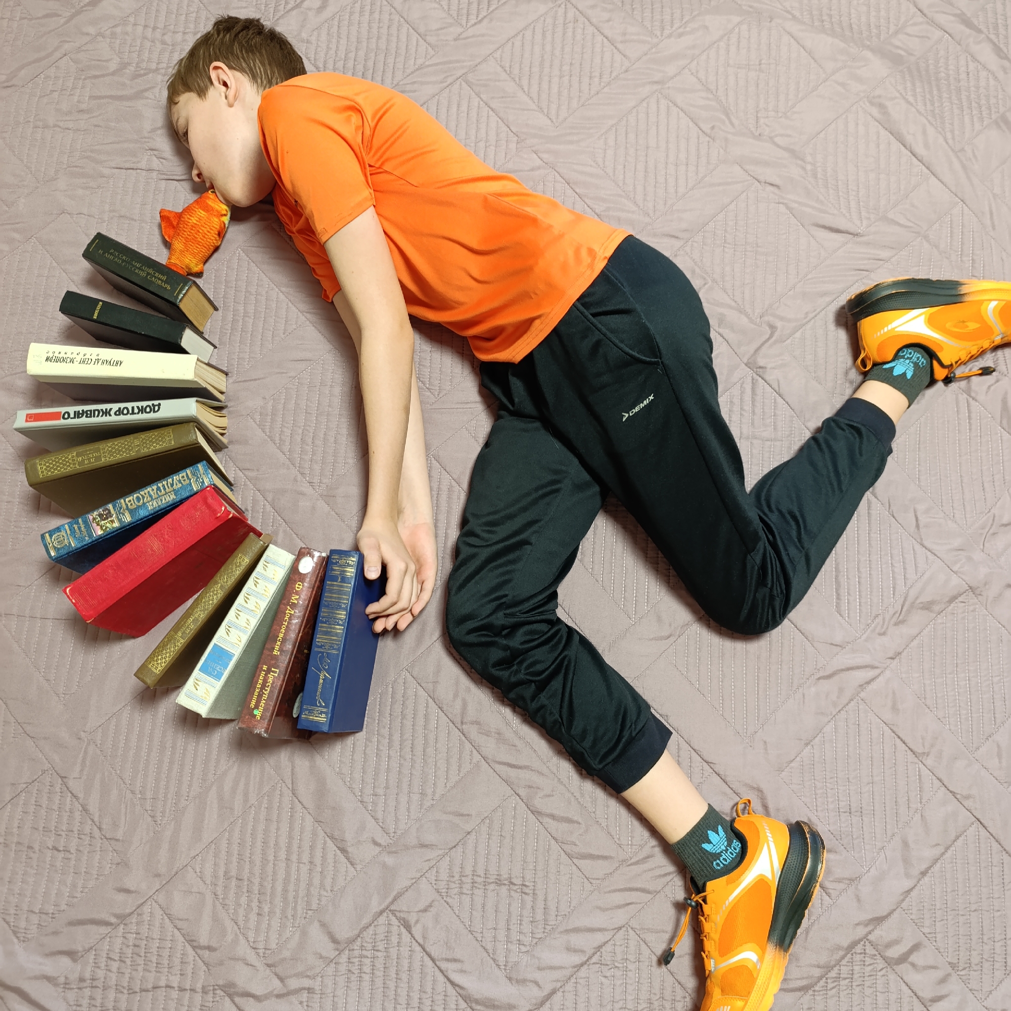 A person wearing an orange t-shirt and black sweatpants lies on a bed covered with a brown, quilted blanket. The person's head rests atop a small pile of books and an orange fish-shaped toy. The person's left hand rests on a larger collection of books arranged in a semicircle around their head. They also wear orange running shoes and black socks with the Adidas logo. The books are in Russian and include titles such as "Doctor Zhivago", "Crime and Punishment", and "Russian-English and English-Russian Dictionary". The sweatpants have the "Demix" logo. The location is possibly in the city of Марковский, Russia.