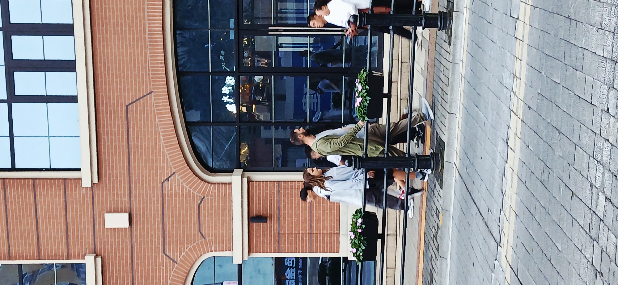 The image captures a group of people in what appears to be the Yuzhong District of Chongqing, China, near a building with brickwork and large, reflective windows. The building has a combination of rectangular and arched window styles. A street or sidewalk runs alongside the building, paved with gray stones. A black iron railing separates the sidewalk from the building, with flower boxes attached to it. The windows reflect Chinese characters in neon lighting, suggesting the presence of nearby establishments. The people seem to be walking or standing near the railing, and their clothing is varied.
