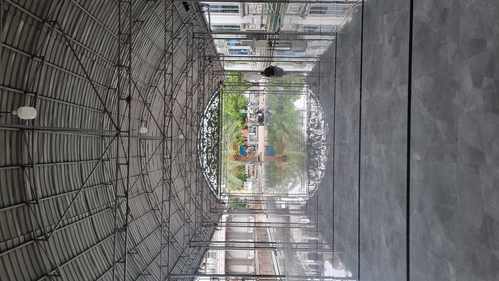 The image depicts a large, open-air covered walkway or pavilion with a corrugated metal roof supported by a metal frame structure. The floor is paved with large, reflective grey tiles, showing a clear reflection of the roof and the surroundings.
In the middle ground, there is a prominent palm tree, a few parked vehicles including what appears to be a scooter or motorcycle, and some other objects that are difficult to discern clearly. Beyond this, there are buildings with architectural details, greenery, and a few more vehicles.
On the right side of the walkway, a person is seated on the floor, facing away from the camera and towards the outside. The person is wearing dark clothing.
The scene is located in Keudekruenggeukueh, Indonesia, and the overall environment suggests a public or communal space, possibly associated with a religious site or a gathering area. The lighting indicates it is daytime, and the reflective floor suggests it may have been recently cleaned or rained upon.