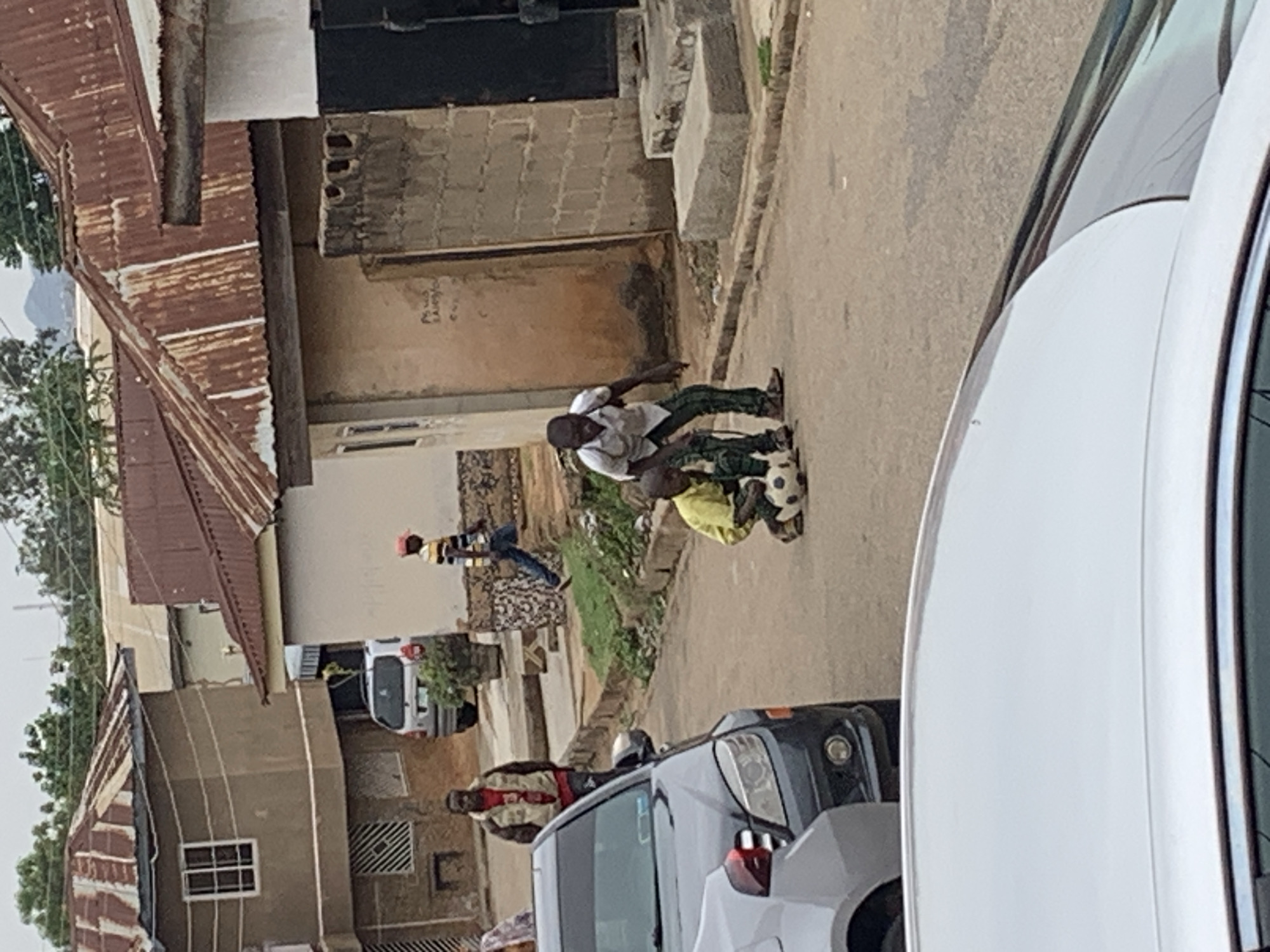 The image shows a street scene in Jos, Nigeria. Two children are wrestling over a soccer ball in the middle of the road. One child is wearing a white shirt and green pants, while the other is wearing a yellow shirt. A man wearing a striped shirt and vest stands next to a dark-colored SUV. A white car is partially visible in the foreground. Several buildings with corrugated metal roofs line the street. In the background, other people are walking and a white vehicle is parked.