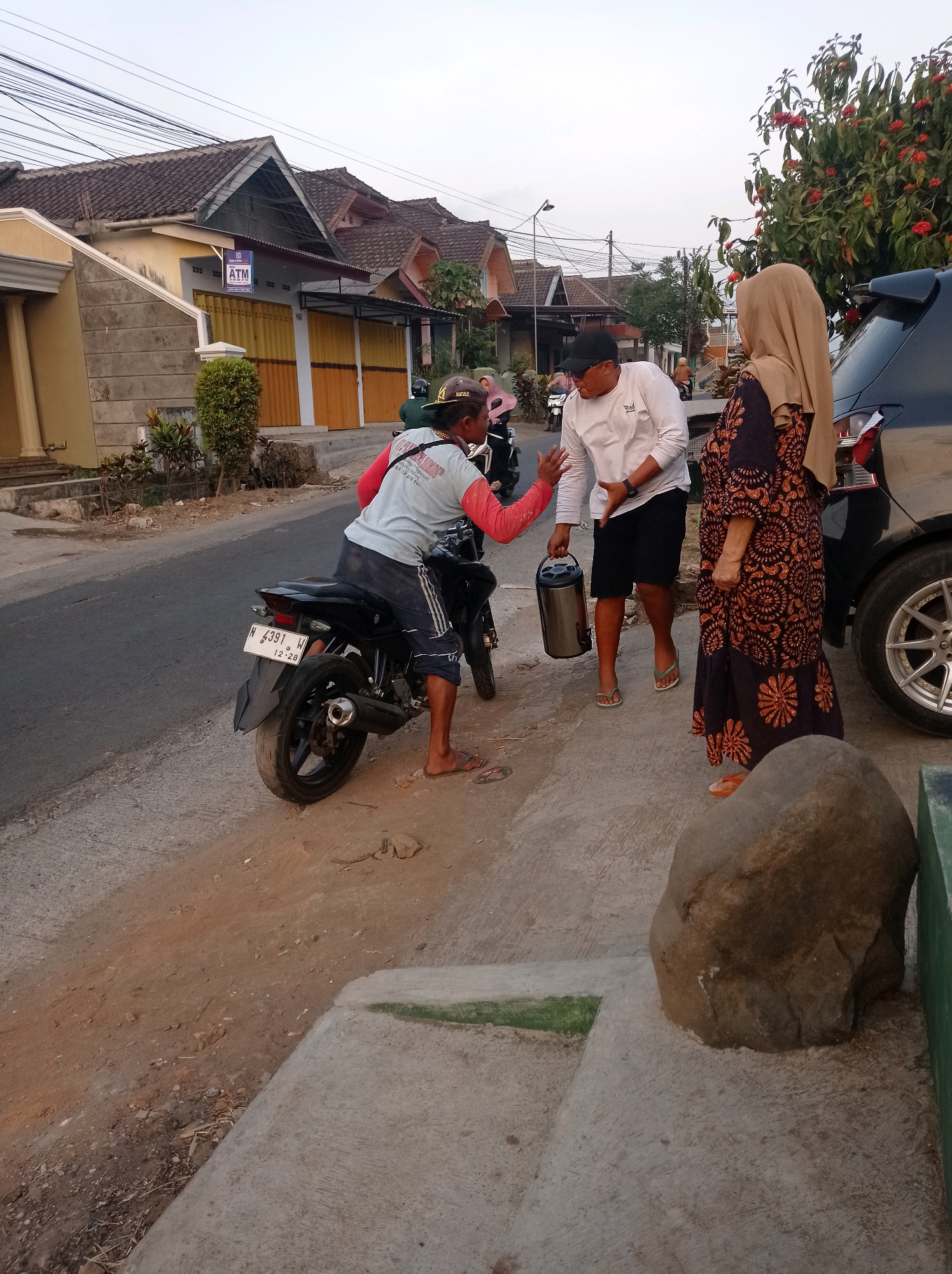 In Purwosekar, Indonesia, a scene unfolds on a residential street at dusk. Three individuals are engaged in an interaction near a parked motorcycle and a black SUV. On the left, a person on a black motorcycle, wearing a light blue shirt and dark shorts, gestures with their right hand. In the center, a man in a white long-sleeved shirt and black shorts, holding a metallic cylindrical container, faces the person on the motorcycle. To the right, a woman in a patterned dress and a beige hijab looks towards the man holding the container. The background features a row of houses with tiled roofs and yellow and orange roller shutters, a streetlamp, and overhead power lines. The ground is a mix of asphalt, dirt, and concrete, with a large boulder near the foreground.
