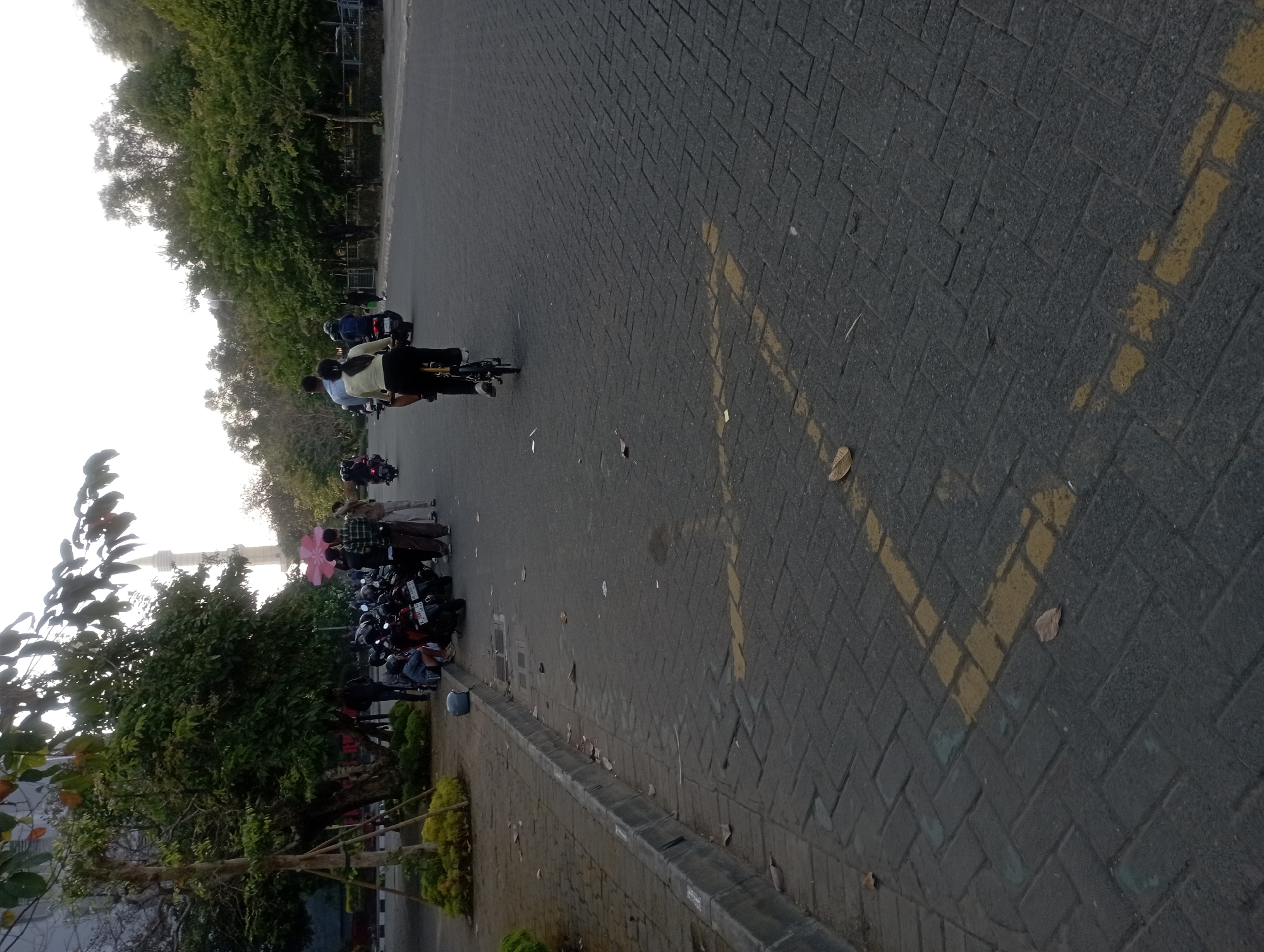 This is a medium shot, taken from a low angle, of a paved area in Depok, Indonesia, likely a parking lot or walkway adjacent to a street. In the foreground, a gray paved surface with yellow markings is visible. The yellow markings appear to be a dashed line and a cross. Scattered leaves are on the ground. To the left, a gray curb separates the paved area from a sidewalk or landscaped area with plants and trees. A tall, thin structure, resembling a minaret, is visible in the background, suggesting the location might be near a mosque. Several motorcycles are parked on the left side of the image, and a few people are standing near them. In the middle ground, a person is riding a bicycle on the paved surface, with another person riding behind them. Further in the background, more trees and buildings are visible. The sky is overcast, and the lighting suggests it is either early morning or late afternoon.