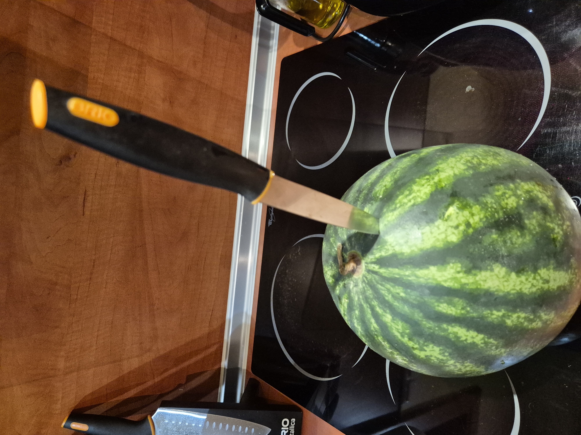 A close-up, eye-level shot depicts a whole watermelon on a black, glass-top electric stove. A knife is embedded in the top of the watermelon, its blade extending partially into the fruit. The watermelon exhibits its characteristic green rind with darker stripes. The stove features two white circular heating elements, one of which is partially obscured by the watermelon. In the background, a wooden-textured countertop and cabinetry are visible. To the left, a portion of another knife handle and packaging are present. A bottle of olive oil is visible to the right. The scene appears to be indoors, likely in a kitchen.