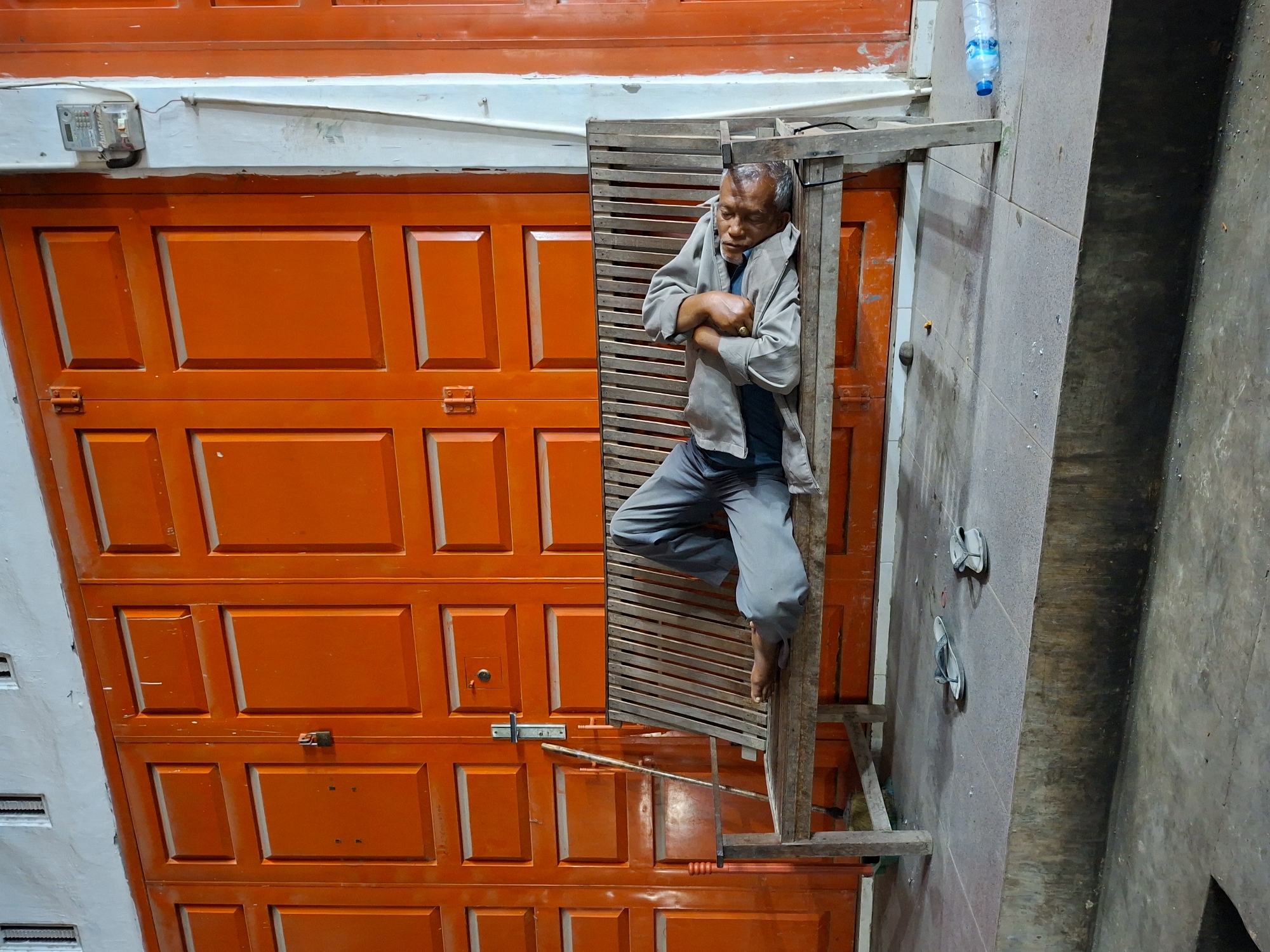 The image depicts an individual resting against a makeshift wooden structure, appearing to be a scaffold, set against a bright orange paneled door. The scene is located in Keudegeudong, Indonesia, based on available location data.
The individual is an older male, dressed in gray pants, a dark t-shirt, and a gray jacket. He is leaning against the wooden scaffold, with his arms crossed. His expression appears relaxed, suggesting he may be sleeping or resting.
The scaffold is constructed of rough-hewn wooden planks, appearing utilitarian and somewhat unstable. A clear plastic water bottle sits atop the scaffold.
The bright orange door features a paneled design. A metal rod, possibly for locking the door, is visible. A pair of flip-flops are positioned on the ground beside the scaffold.
The environment suggests an urban setting, with a gray concrete wall and floor visible on the right side of the frame.
