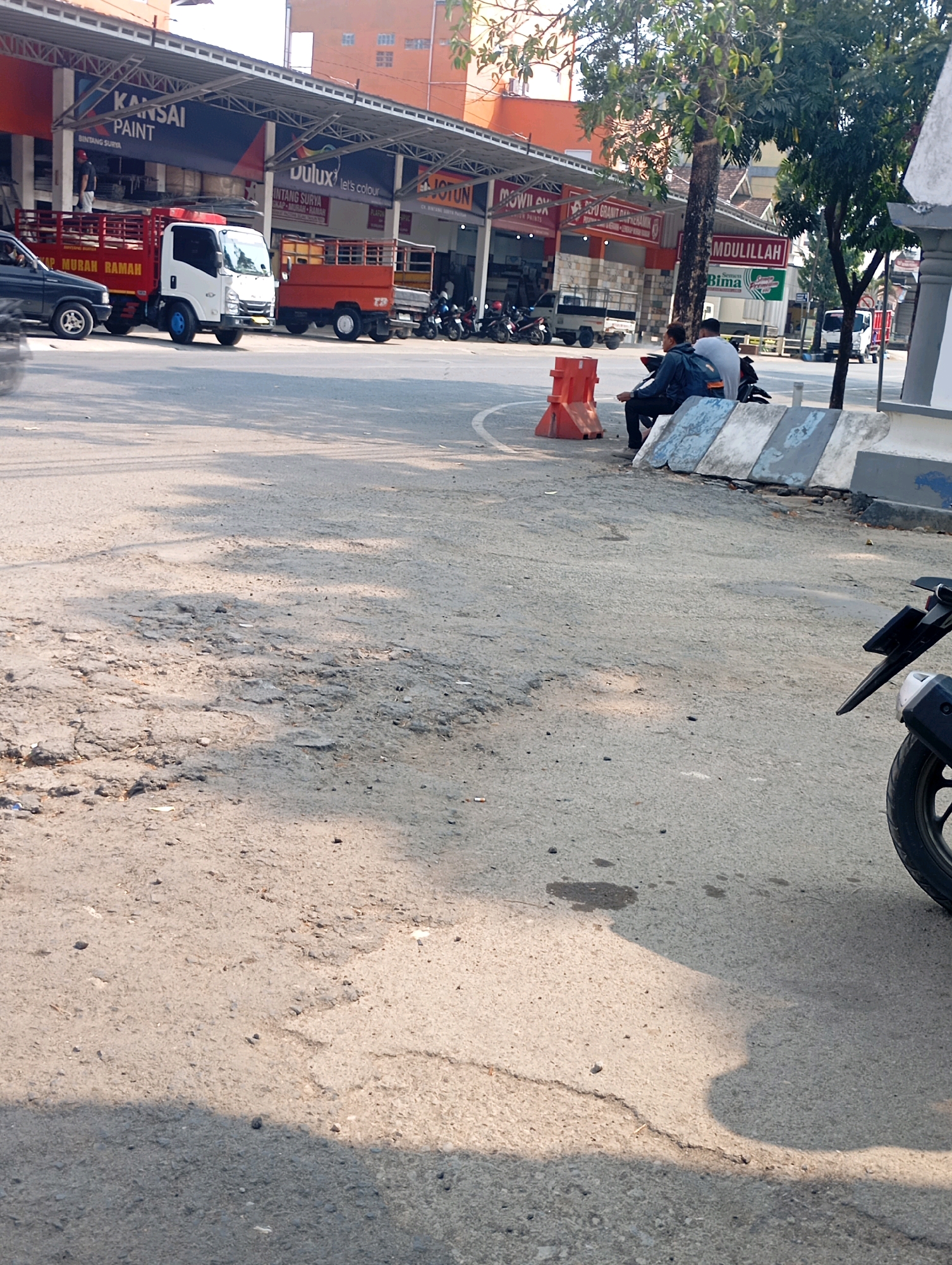 The image depicts a street scene in Pacitan, Indonesia, likely near a commercial area. In the foreground, a cracked asphalt road shows shadows from trees and overhead structures, indicating it is daytime. To the left, a white and red truck is parked, with a dark blue car behind it. Further down the street, an orange truck is also parked. In the middle ground, two individuals are seated on a motorcycle on the side of the road, facing away from the camera. They are near orange traffic barriers and concrete dividers. The background features buildings with awnings and signage, including "KANSAI PAINT" and "DULUX". Trees are visible, contributing to the urban environment. The overall impression is a moderately busy street with commercial activity and parked vehicles.