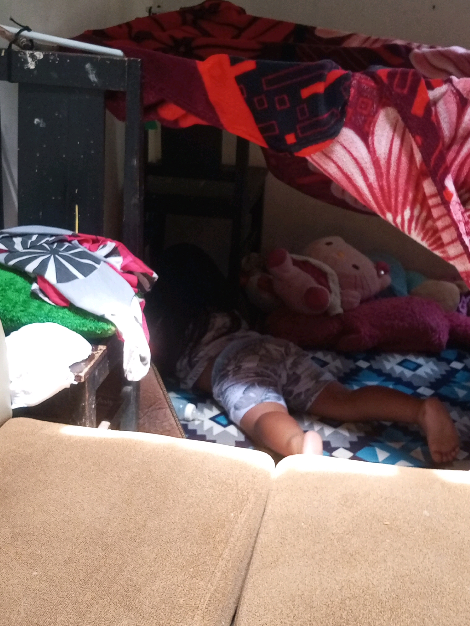 In Krasak, Indonesia, a child is lying down, seemingly asleep, amidst a makeshift fort constructed from blankets and bedding. The fort is situated inside a room, with various blankets draped to create the enclosed space. A small table with some items, including a green cloth and a patterned fabric, is visible to the left. The child is wearing patterned shorts and is positioned on a patterned bedsheet. Several plush toys are also present within the fort, including a pink Hello Kitty toy and a fuzzy pink stuffed animal. The overall environment suggests a domestic setting, possibly a bedroom, where a child has created a play area.