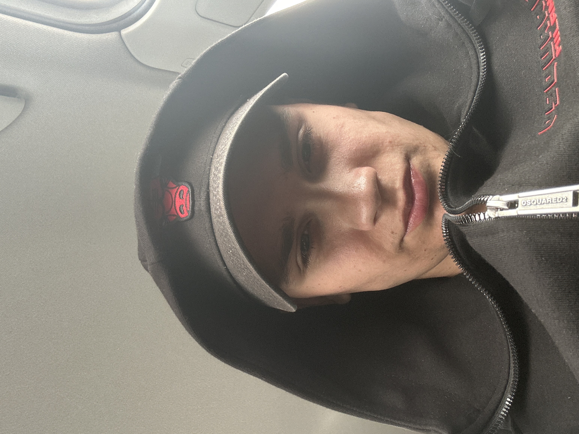 A person is in what appears to be the interior of a vehicle. The person has a dark hoodie with a zipper opened to the upper chest, revealing skin and a sliver of a red lip. On the hoodie's zipper pull tab, the word "DSQUARED2" is visible. The person is also wearing a grey cap with a red emblem, partially obscured by the hoodie. The background shows the interior of the car. The exact location in Sofia, Bulgaria, cannot be determined from the image.