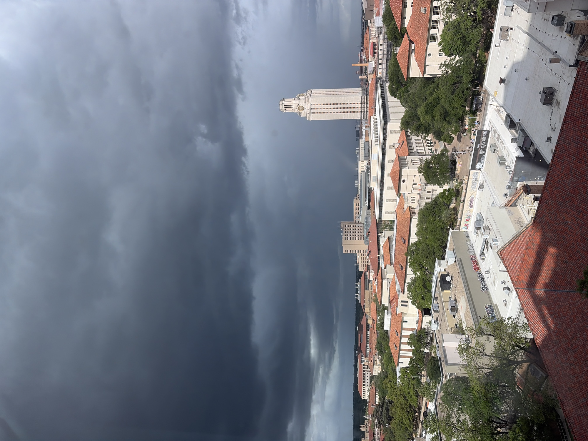 This is a vertically oriented aerial view of the University of Texas at Austin campus in the city of Austin, United States of America. A storm front occupies the left side of the frame. The iconic UT Tower is centered in the frame and rises above the campus buildings. The campus architecture features buildings with red-tiled roofs. Trees are scattered throughout the campus. The buildings are densely packed.