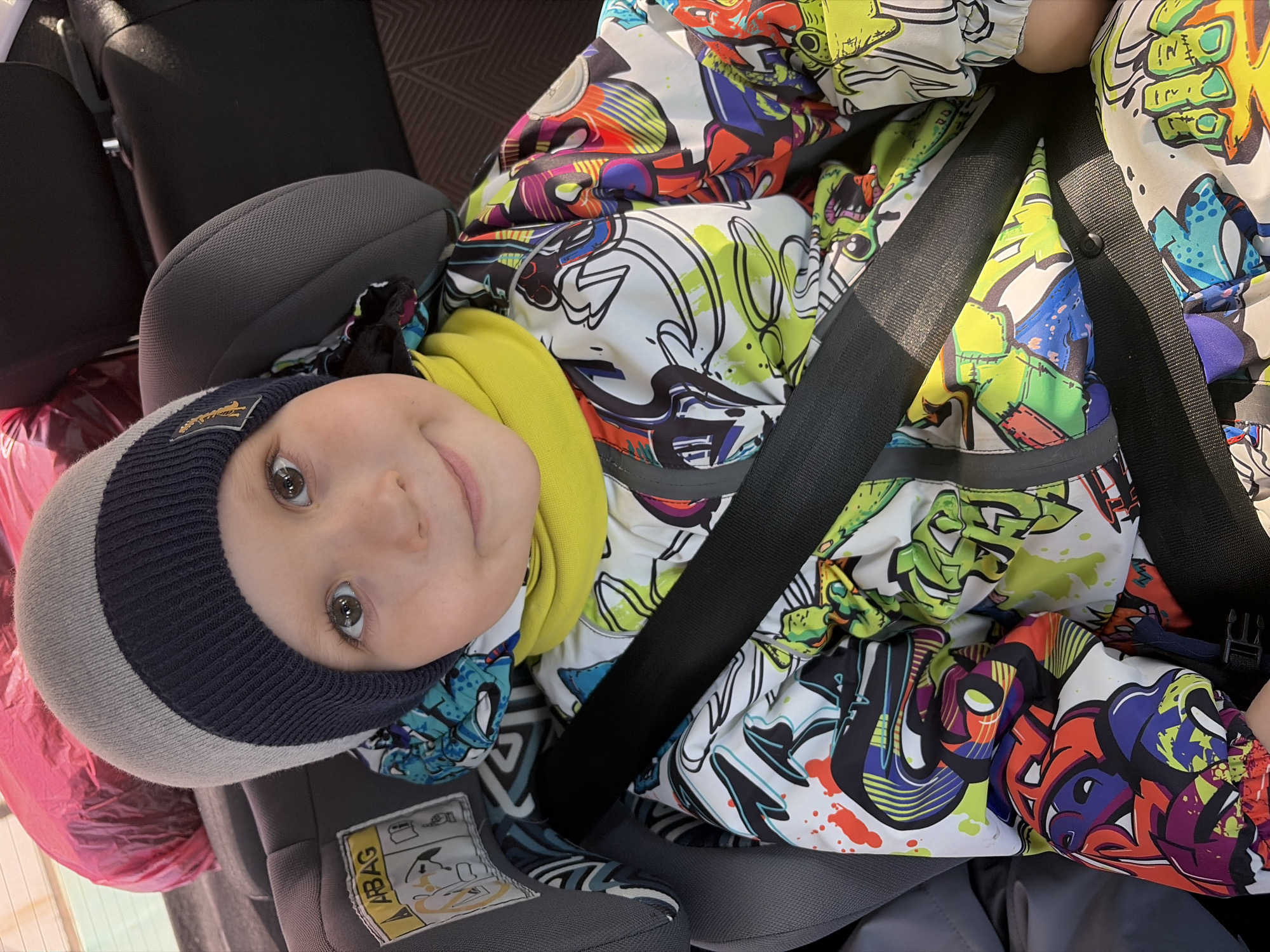 A young child is securely fastened in a grey car seat with a black seatbelt. The child wears a navy and grey striped beanie, a bright yellow turtleneck, and a colorful jacket with graffiti-style designs. The seat has a sticker on the side that says "ABAG." A pink bag is visible in the background. The location is unknown.
