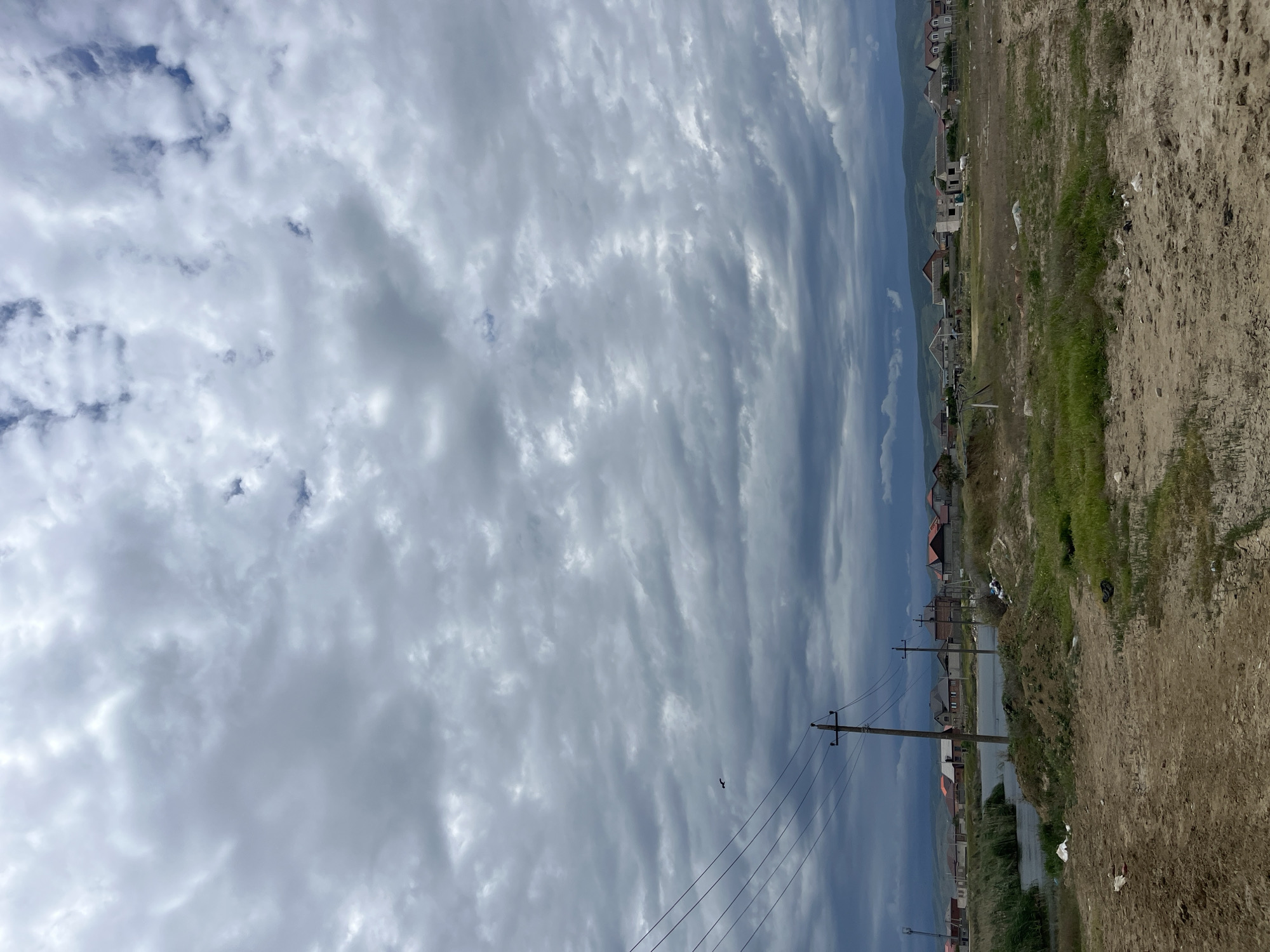 The image shows a view in Kaspiysk, Russia. The sky is filled with textured, gray clouds. A bird is visible in the sky near a utility pole with power lines. The ground is a mix of dirt and sparse green vegetation, suggesting an undeveloped or semi-rural area. In the distance, there are houses and possibly buildings along a waterline, indicating a coastal or waterside location. There are no people visible in the scene. The lighting suggests an overcast day.