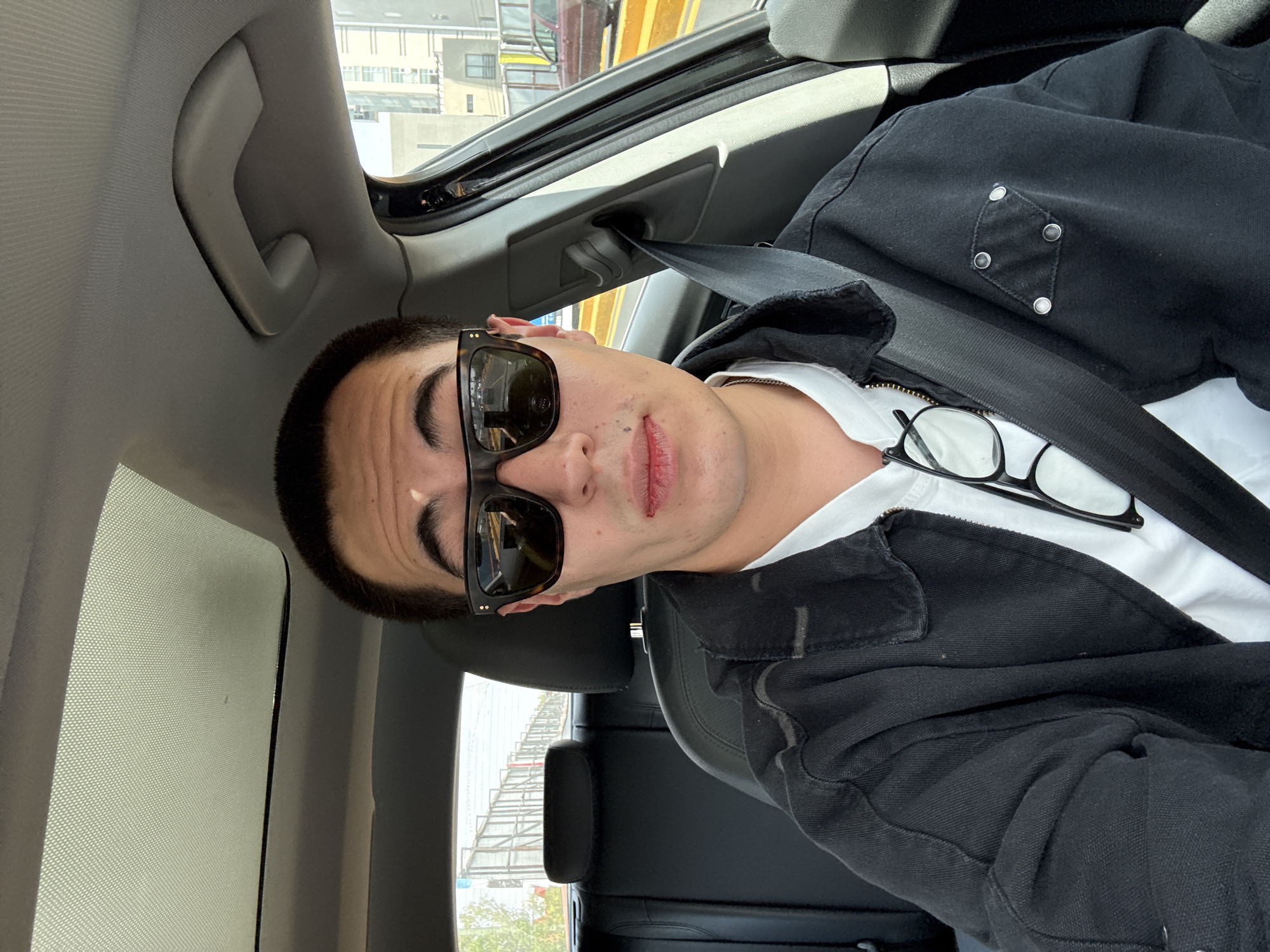A young man is captured in a selfie or portrait, seated in the passenger side of a car during what appears to be a sunny day in Puebla, Mexico. He is looking directly at the camera with a neutral expression, his lips slightly parted. He has short dark hair and prominent eyebrows, and is wearing dark rectangular sunglasses that obscure his eyes.
He is dressed in a white t-shirt layered under a black denim or work-style jacket. The jacket features distinct silver stud or rivet detailing in a diamond pattern on the left side of the chest. A pair of clear-framed eyeglasses is tucked into the collar of his white t-shirt. A black seatbelt crosses his chest.
The car's interior is visible, featuring a light grey overhead grab handle on the roofline and a dark grey or black headrest from the seat behind him. Above, a sunroof or panoramic roof is partially covered by a textured, light-colored sunshade, allowing dappled light into the vehicle.
Outside the car, an urban landscape unfolds, with modern buildings and what appears to be an elevated structure or scaffolding with prominent yellow and red elements, suggesting an industrial or construction area. The bright daylight creates clear shadows and highlights the vehicle's interior. There is no visible text in the image.