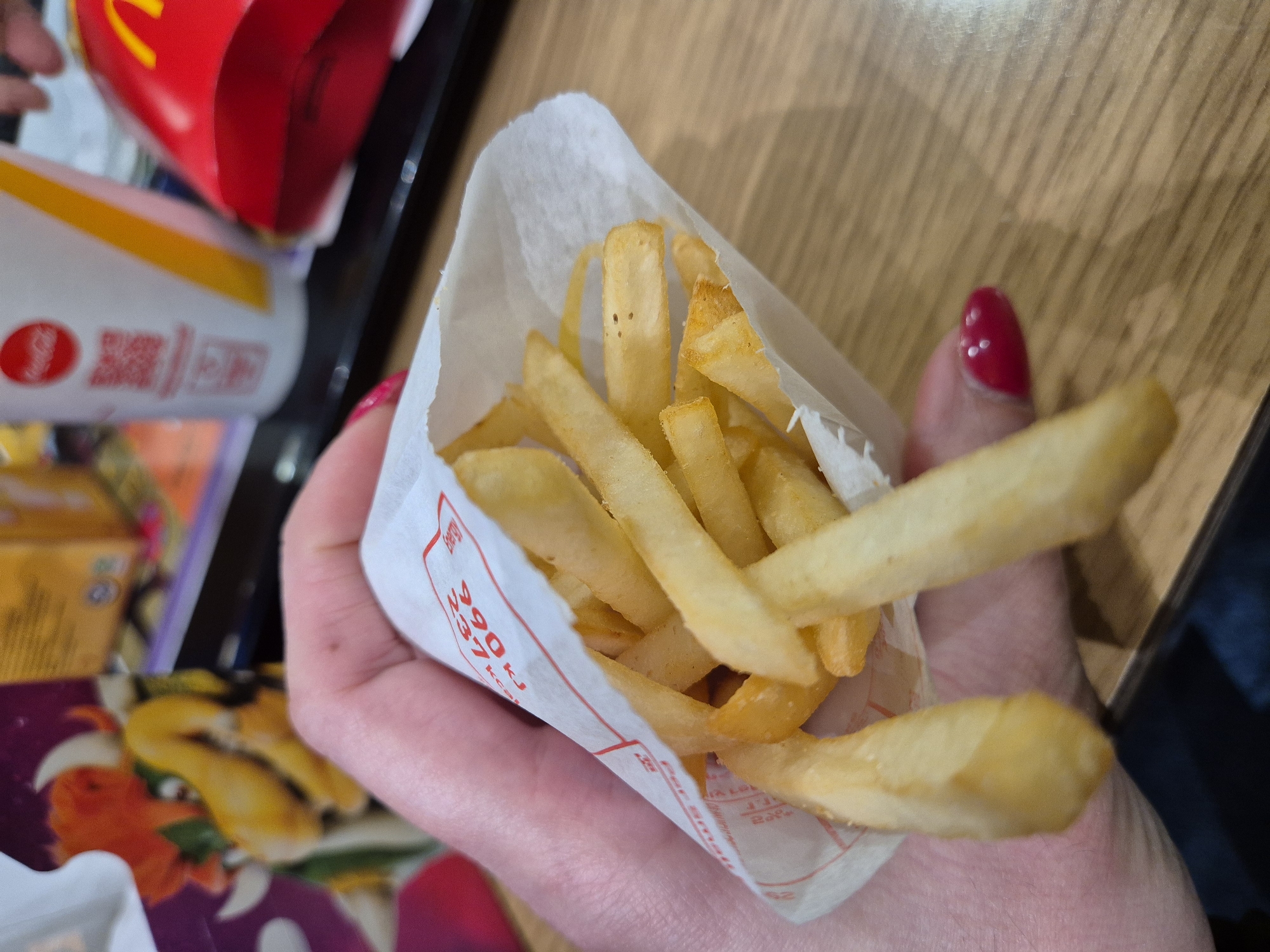 A person holds a small bag of McDonald's french fries. The bag is white with red lettering indicating nutritional information. Two fries extend out from the bag. The person's hand has a red fingernail. In the background, a McDonald's drink cup and other McDonald's packaging are visible. The scene takes place at a table, with the table surface being a light-colored wood grain. The precise location within West Suffolk, United Kingdom cannot be determined from the image.
