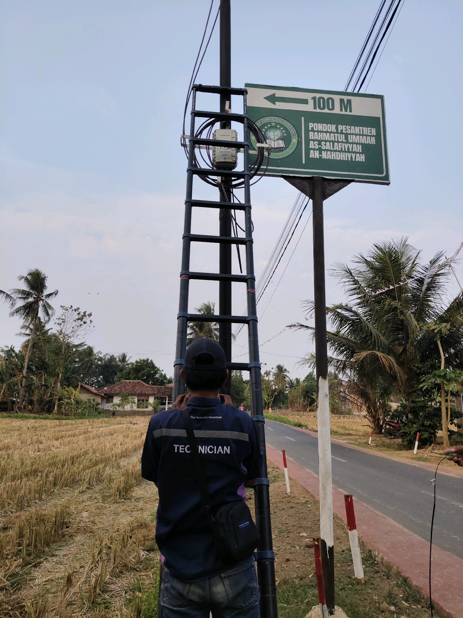 The image depicts a technician in Sidosari, Indonesia, standing next to a utility pole. The technician is wearing a dark blue shirt with "TECHNICIAN" printed on the back and jeans. They are holding what appears to be a cable and looking upwards towards equipment on the pole. A signpost indicates "100 M" with an arrow pointing left, and text in Indonesian that translates to "Pondok Pesantren Rahmatul Ummah As-Salafiyyah An-Nahdhiyyah," signifying a pesantren or Islamic boarding school. The environment is rural, with a harvested rice field to the left, palm trees, other vegetation, and a road to the right. The sky is clear and blue. The utility pole has a ladder attached and various electrical equipment mounted on it.
