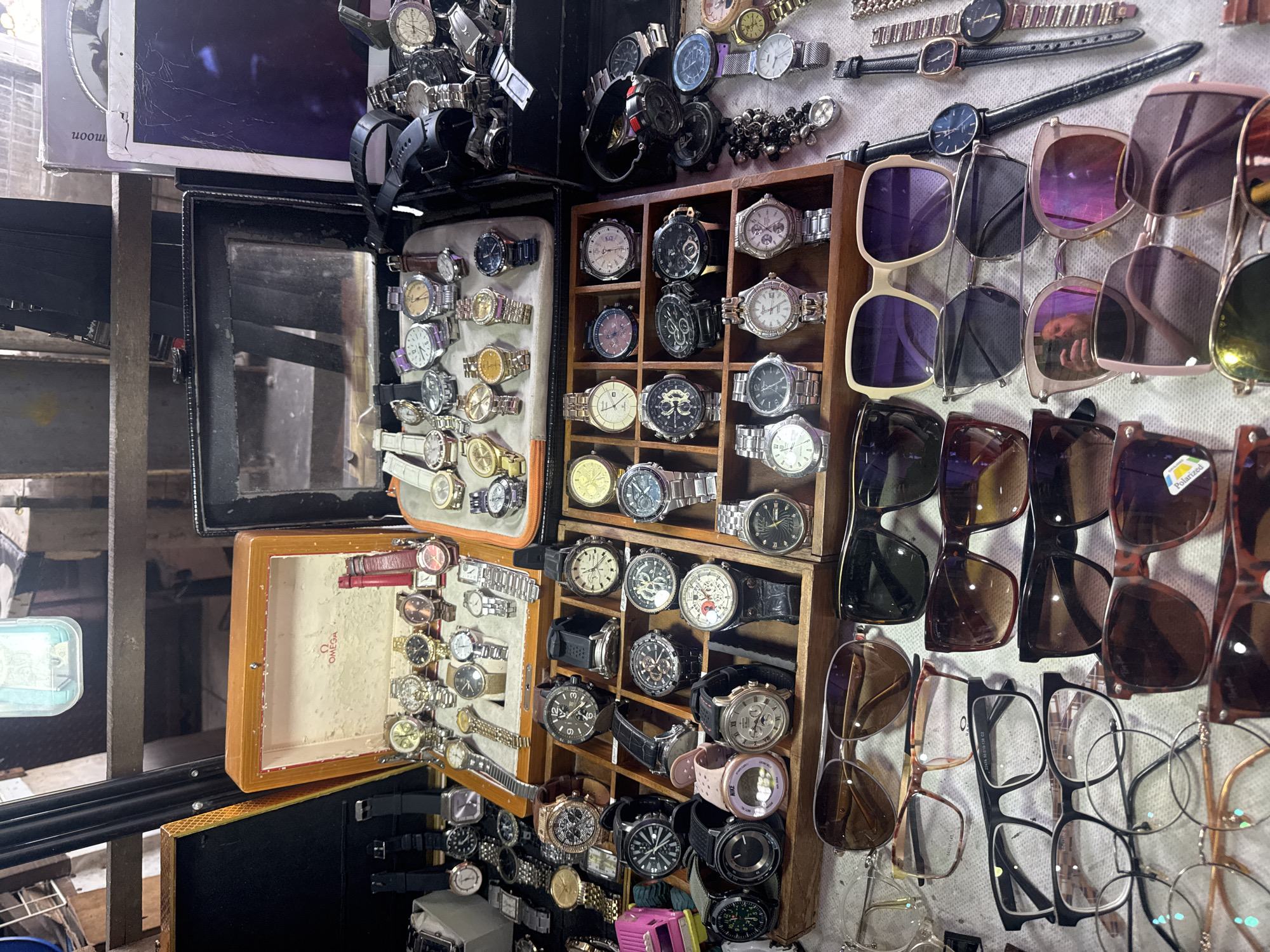This image shows a display of watches and sunglasses in a market stall. The watches are laid out in wooden trays, with some in an open box that says "OMEGA." The sunglasses are stacked below the watches, with various styles and colors visible. The background shows shelves filled with more items, suggesting a busy marketplace. The overall impression is of a vendor selling accessories.