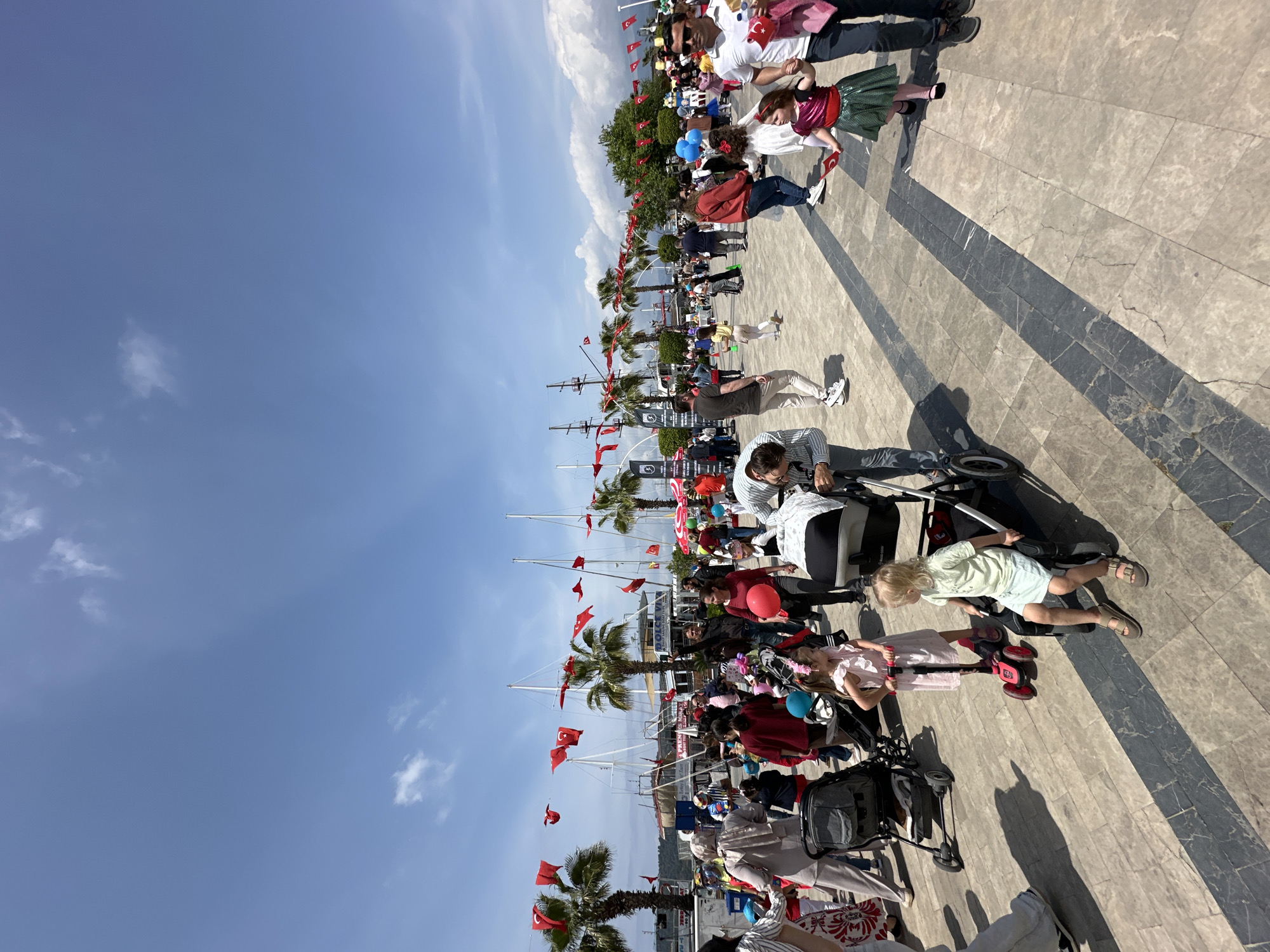 The image depicts a sunny day at the Fethiye waterfront in Turkey. A crowd of people, including families with children in strollers, are gathered along a wide, patterned stone walkway. Many Turkish flags are visible, affixed to flagpoles and held by individuals, suggesting a national celebration or event. Palm trees line the walkway near the waterfront, where several boats are docked. The sky is clear and blue, providing a bright backdrop to the lively scene.