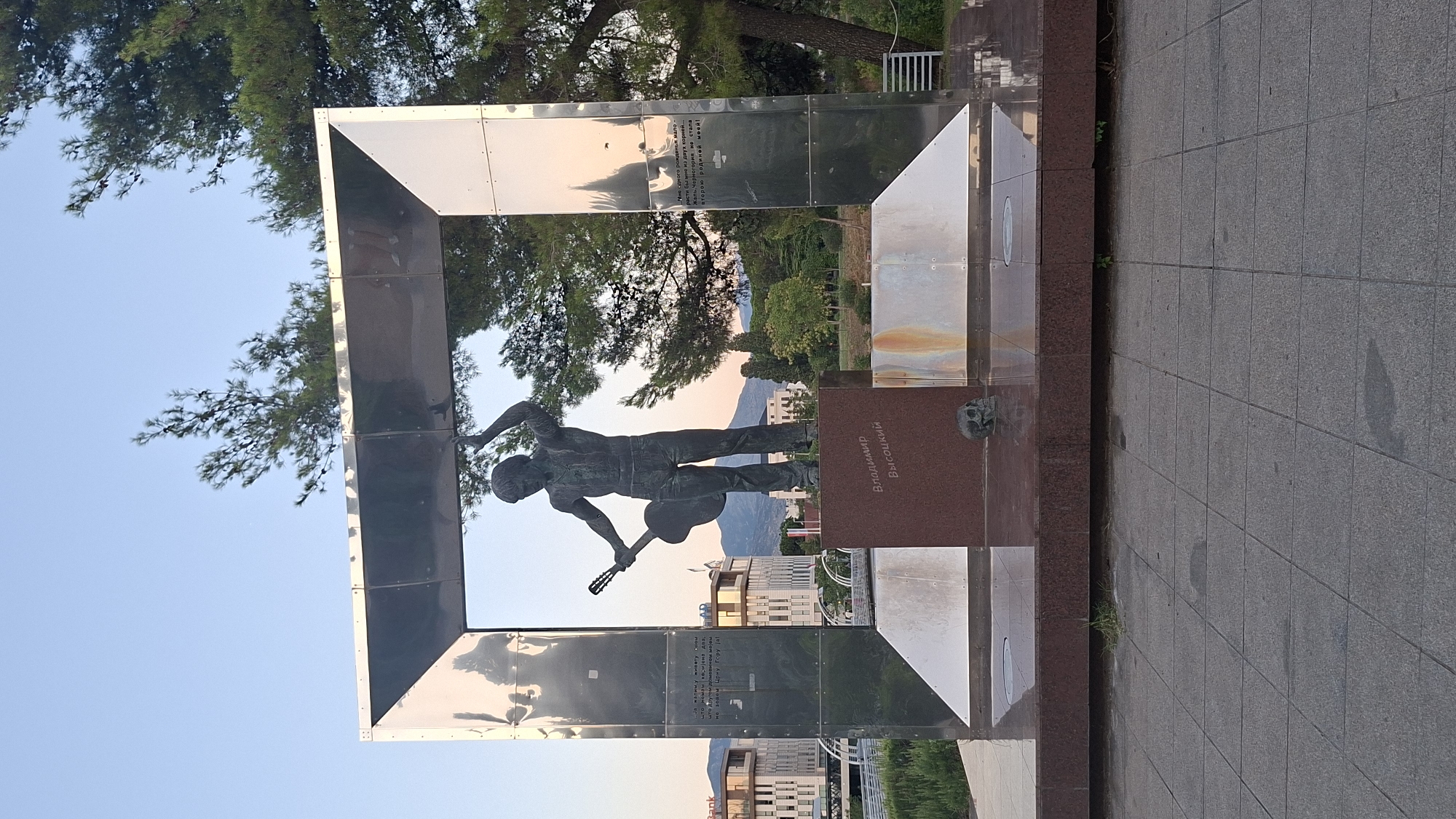 The image shows a monument of Vladimir Vysotsky in the city of Podgorica, Montenegro. The monument features a bronze statue of Vysotsky, a Soviet singer-songwriter, poet, and actor, holding a guitar. The statue is placed on a red granite pedestal inscribed with his name in Cyrillic. To the left and right of the statue are large, polished metal panels, forming a frame around the monument. On the pedestal, at the base of the statue, there is a small, dark skull sculpture. Behind the monument, there are modern buildings, trees, and hills in the background. The sky is clear and blue, indicating daylight. The ground in front of the monument is paved with square tiles.