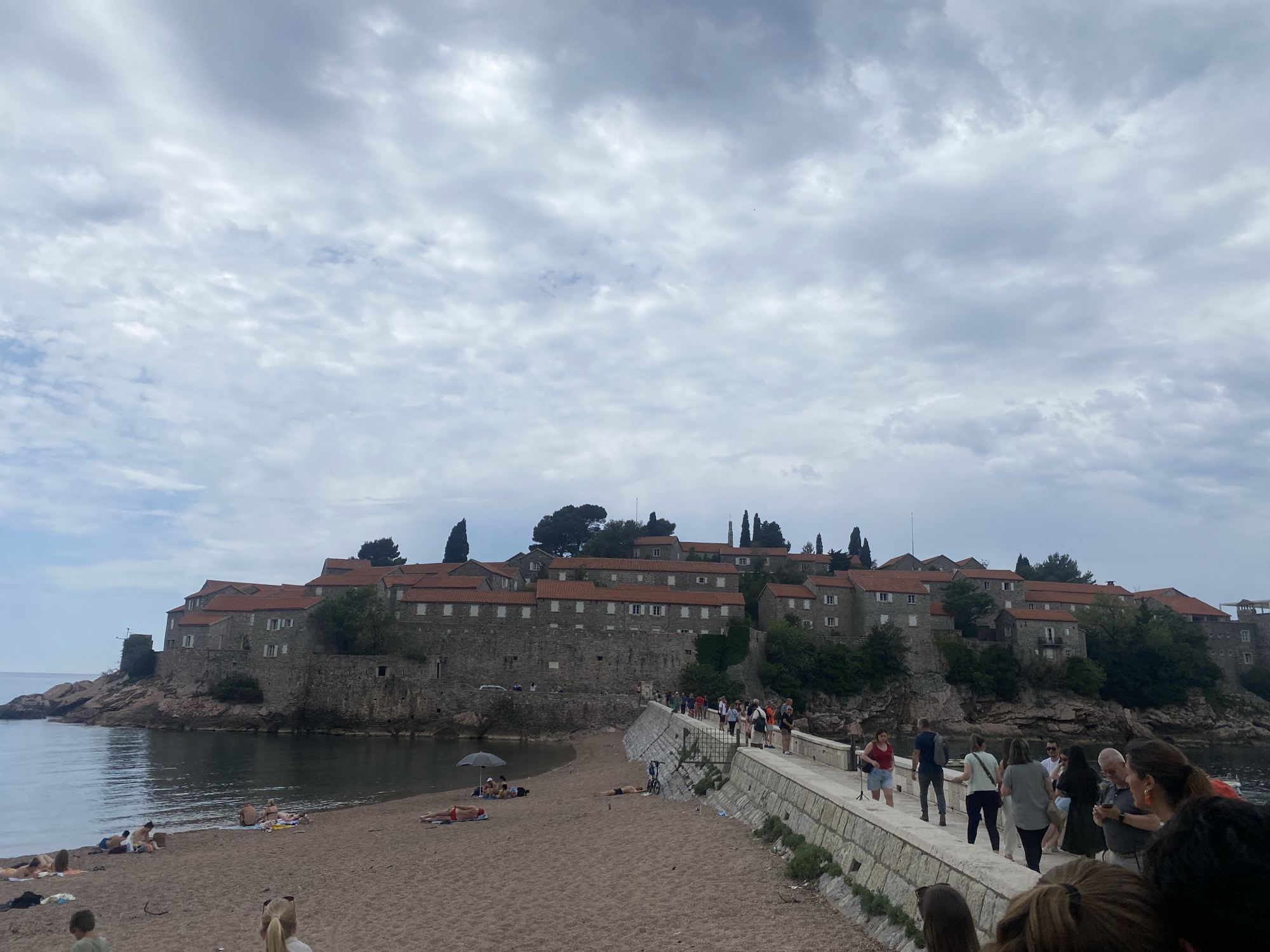 The image shows the island city of Sveti Stefan, Montenegro, viewed from the mainland across a narrow isthmus. The city is built of stone buildings with orange tile roofs. The sky is overcast. On the mainland side, a sandy beach is visible with people sunbathing. A concrete walkway with stone borders is populated by numerous pedestrians.