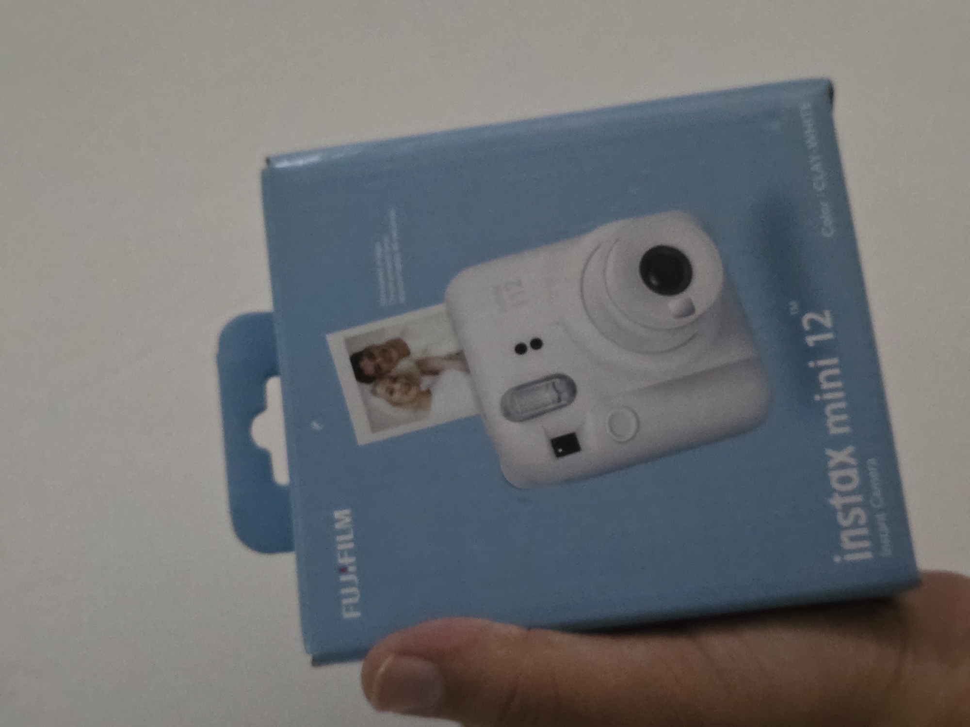 A hand holding a light blue box with "FUJI.FILM" written in white letters across the top. A smaller image of a couple is printed on the front of the box, and a white instant camera with a black lens is depicted. The camera is labeled "instax mini 12". Below the camera illustration, the words "instax mini 12" and "Instant Camera" are printed in white. To the right, "Color: CLAY WHITE" is written in smaller font. The background is a plain white wall. This image is not located in Qaratau, Kazakhstan.