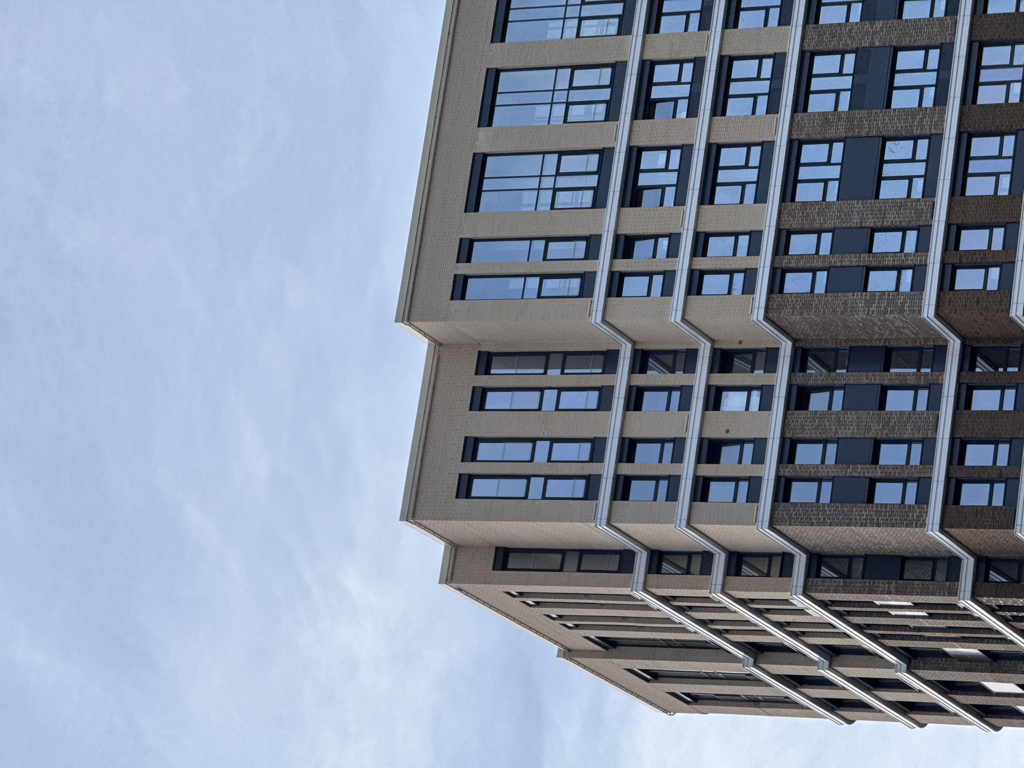 The image shows a tall, modern building viewed from a low angle, looking upwards. The building has a boxy, stepped facade with multiple rows of windows. The exterior is primarily a light tan or beige color with darker brick or paneling sections. Silver-colored trim runs vertically along the building's facade. The sky is visible in the background, pale blue with wispy clouds. There are no people visible. The building is located in Perm, Russia.