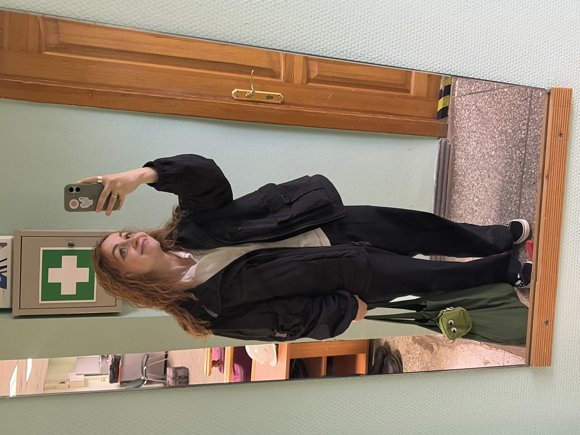 A young woman with long, wavy, reddish-brown hair is depicted taking a mirror selfie. She is holding an olive green smartphone with stickers on its case, raising her left hand to capture her reflection. The woman is dressed in a black zip-up jacket over a white undershirt, black pants, and black sneakers with white soles. A dark green tote bag, featuring a small green pouch with googly eyes, rests at her feet. She casts a soft, upward gaze with a gentle smile, conveying a pleasant expression.
The mirror, framed by light-colored wood, reflects an indoor setting. Behind the woman's head, a wooden door with a gold coat hook is visible. The wall, to the left of the door and surrounding the mirror, is a light, textured green. Prominently featured on the wall to the left is a grey first aid kit cabinet marked with a universal green cross symbol. The reflected floor appears to be a grey, speckled or terrazzo-like surface. Further reflections in the lower part of the mirror suggest other indoor elements such as chairs and furniture, implying a room or corridor within a building. The even lighting suggests an indoor setting, likely illuminated by artificial light. The overall scene appears to be set in a public or institutional building in Chaikovsky, Russia.