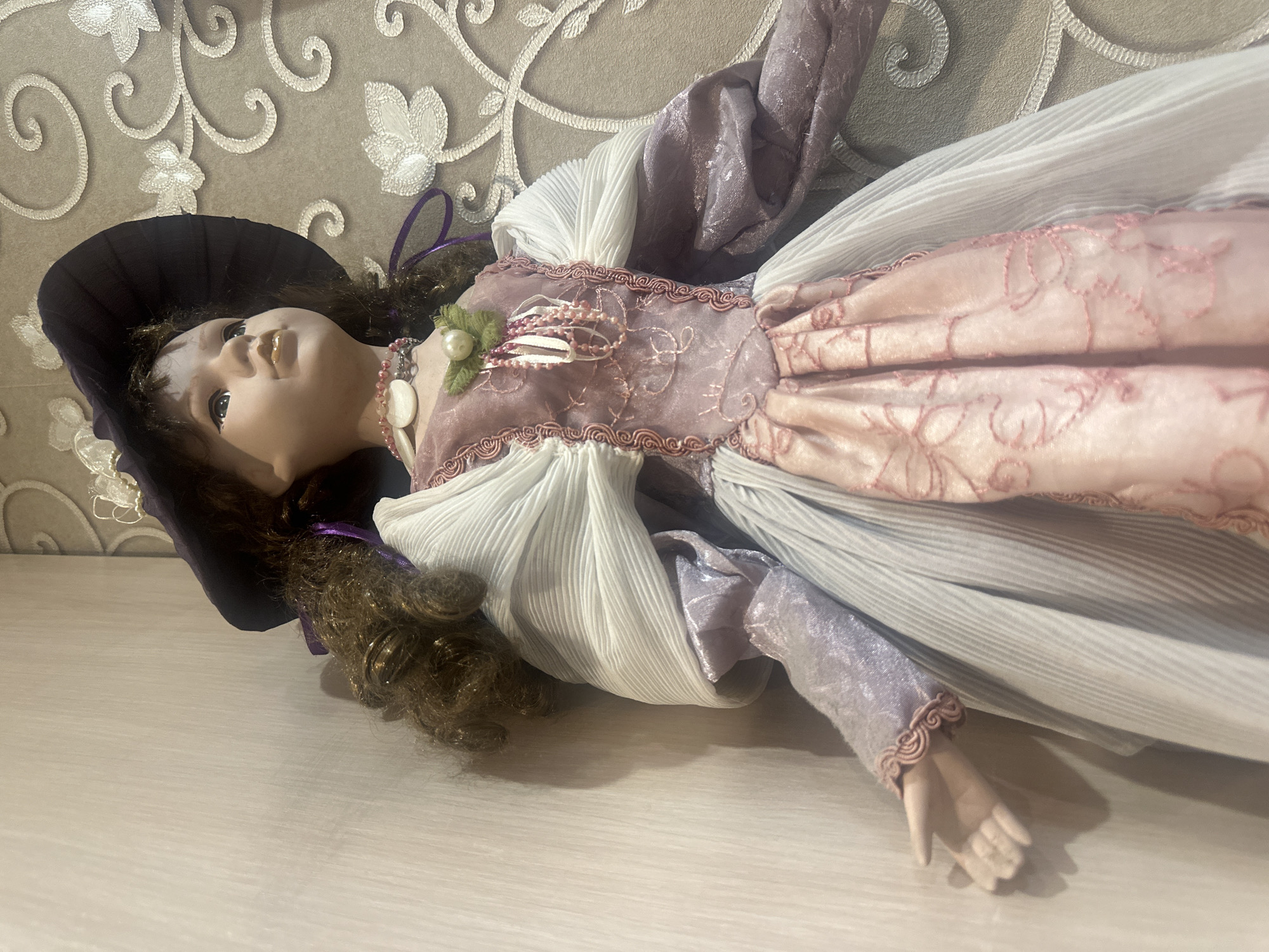 A close-up, eye-level shot shows a porcelain doll dressed in a period-style costume. The doll is positioned in the center of the frame, facing to the right. She wears a wide-brimmed, dark purple hat with purple ribbon accents. Her costume consists of a pale pink bodice with intricate pink embroidery and embellishments, layered over a pale blue and white pleated dress with puffed sleeves. The doll has medium-brown, curly hair that falls past her shoulders. A beaded necklace adorns her neck. The background is a textured wallpaper with a light beige color and a scrolling floral pattern in a lighter, off-white color. To the left of the doll, a plain, light-colored wall is visible, creating a contrast with the patterned background. The lighting is soft, casting a gentle shadow to the left of the doll, suggesting a light source from the right.