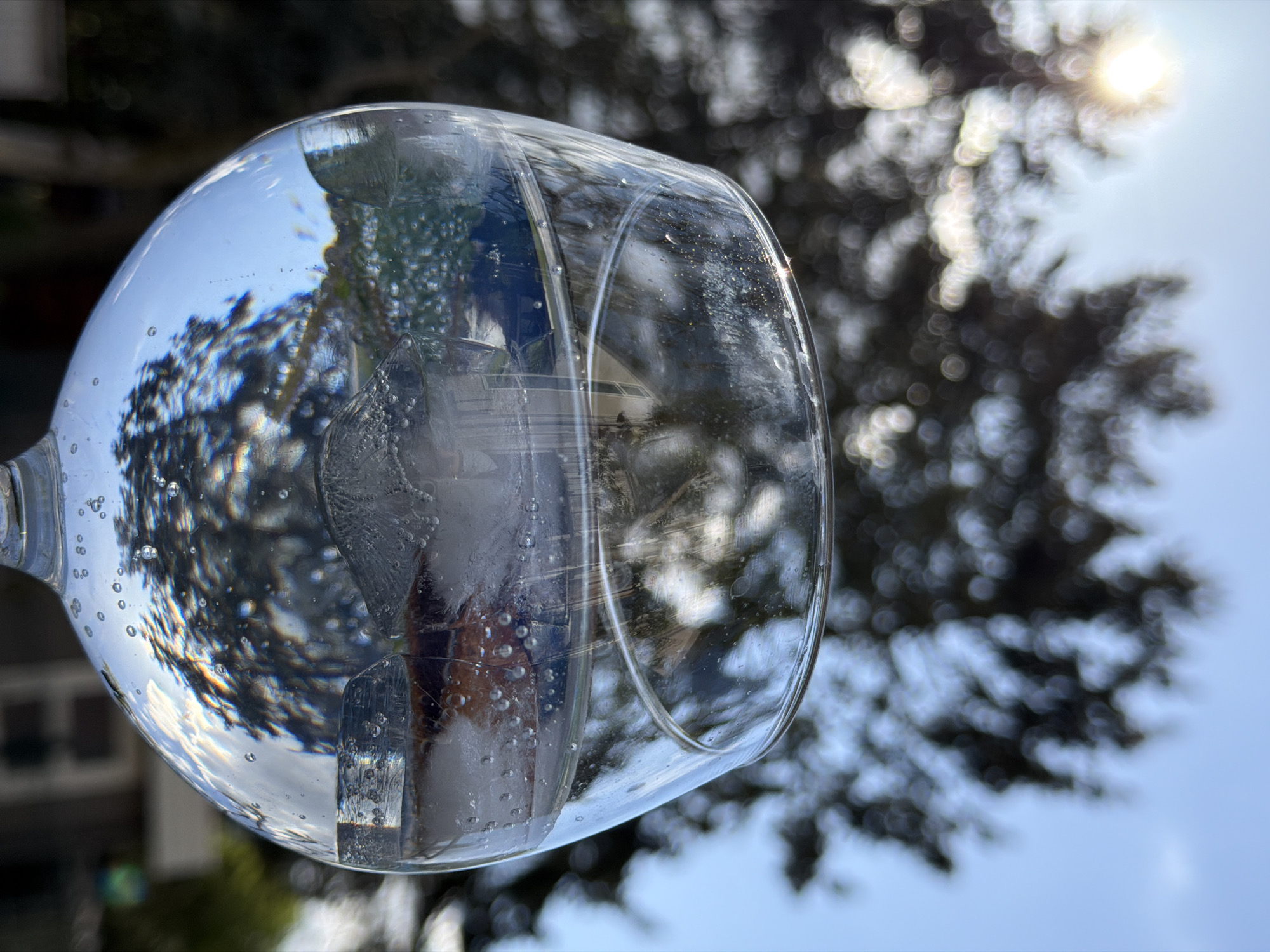 A low-angle, close-up shot shows a wine glass filled with ice and water held against a blurred background of trees and a section of a building. The glass distorts the background, creating an inverted reflection of the scene. Sunlight is visible through the trees. The precise location and time are not discernible from the image alone, but based on visual cues such as building style and common vegetation, it could potentially be in Esch-sur-Alzette, Luxembourg. There is no indication of people or specific actions.