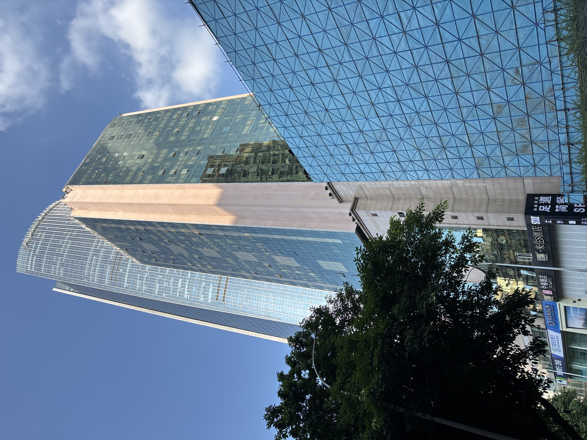 The image shows a modern urban scene in Yuzhong District, China, dominated by towering glass and concrete buildings under a clear blue sky with scattered clouds. In the foreground, a large, leafy green tree partially obscures a streetlamp and the lower portions of the buildings. The central focus is on a skyscraper with a multifaceted glass facade, reflecting the sky and other structures. Adjacent to it is another taller building with a predominantly glass exterior, featuring a light-colored, ribbed vertical element running up its side. To the right, a building is covered in a blue, geometric patterned netting. At the base of the buildings, commercial storefronts are visible, including signage for banks and a hotel. The scene suggests a bustling commercial district with contemporary architecture.