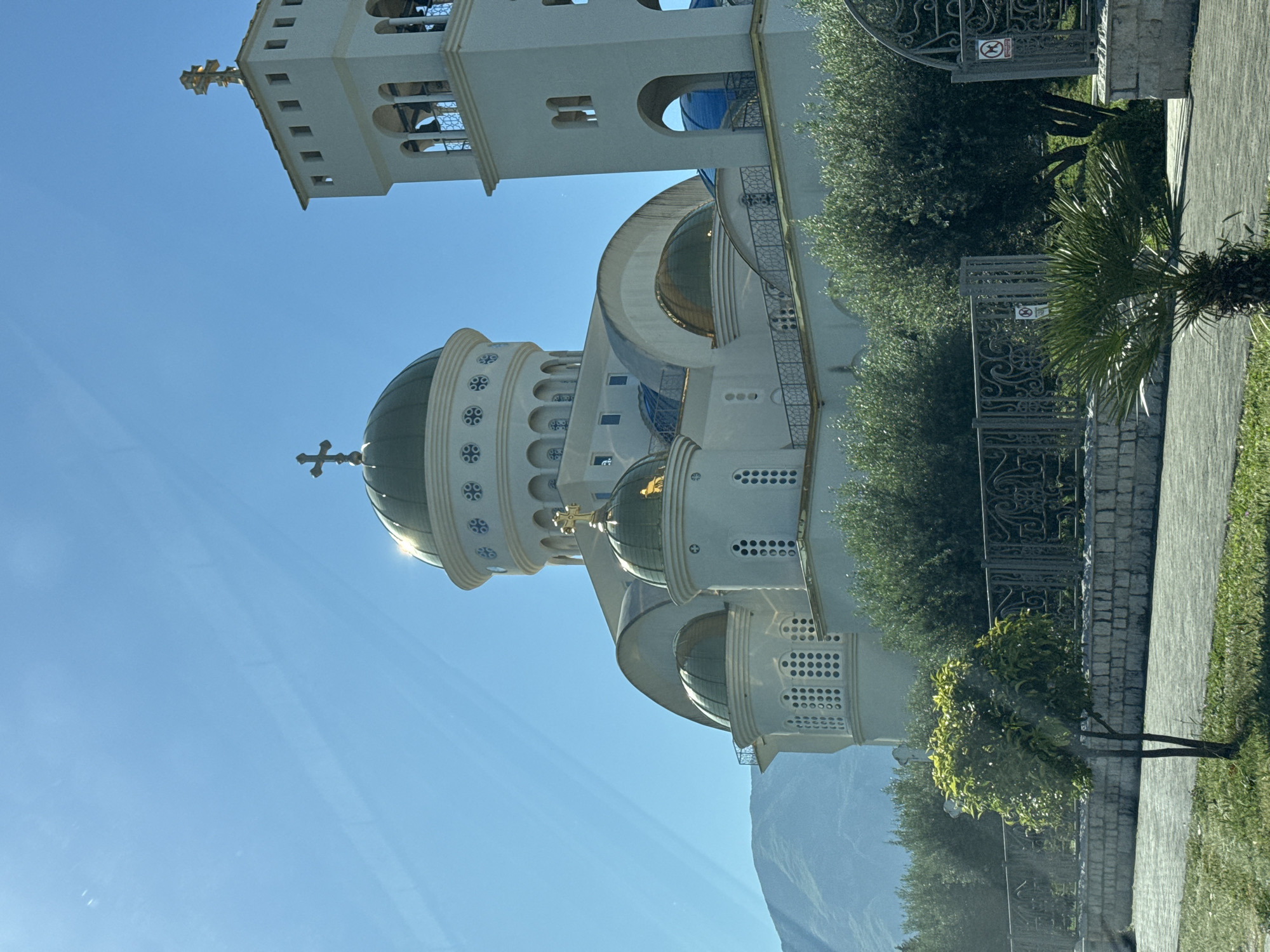 A low-angle shot of the Church of St. John Vladimir in Bar, Montenegro, captured on a sunny day. The architecture features a prominent bell tower with a gold cross at the top, alongside several domes covered in green and gold. The building's facade is white, with architectural details such as arched windows and circular patterns. The church sits behind a wrought-iron fence, with trees and shrubbery visible within the compound. A mountain range appears in the background, partially obscured by the church. The foreground shows a paved pathway and grass. The shot seems to be taken from inside a vehicle, as evidenced by the faint reflections on the window.