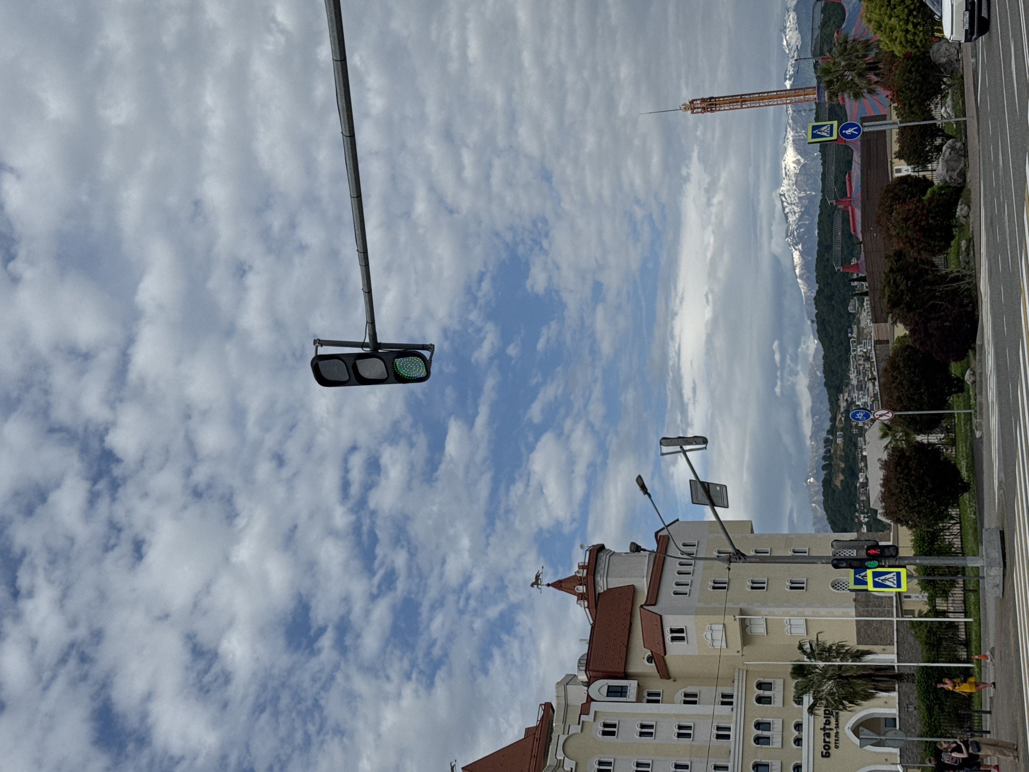 The image shows a street scene in Sirius, Russia, taken on a cloudy day. Dominating the foreground is a multi-story building with a red-tiled roof, light-colored walls, and multiple windows. A weather vane adorns a tower-like structure atop the building. Directly in front of the building are traffic lights displaying a green light in one direction and a red light in another, along with pedestrian crossing signs.
Behind the building, the scenery opens up to reveal a cityscape with buildings and greenery, with snow-capped mountains in the distance under a sky filled with clouds. A tall amusement park ride is visible in the background. A street runs along the right side of the frame, marked with road lines, trees, and shrubbery. People are visible on the street, including a person wearing a yellow shirt. A car is also parked on the street.