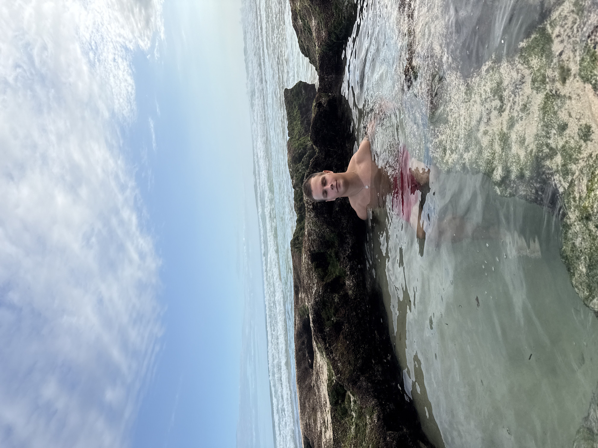 A man is photographed waist-deep in shallow water between dark, mossy rocks. The scene is likely a tide pool in a coastal area. In the distance, a beach is visible, with waves breaking on the shore. Above, the sky is pale blue with scattered clouds. This is potentially located in Kuta Selatan, Indonesia.