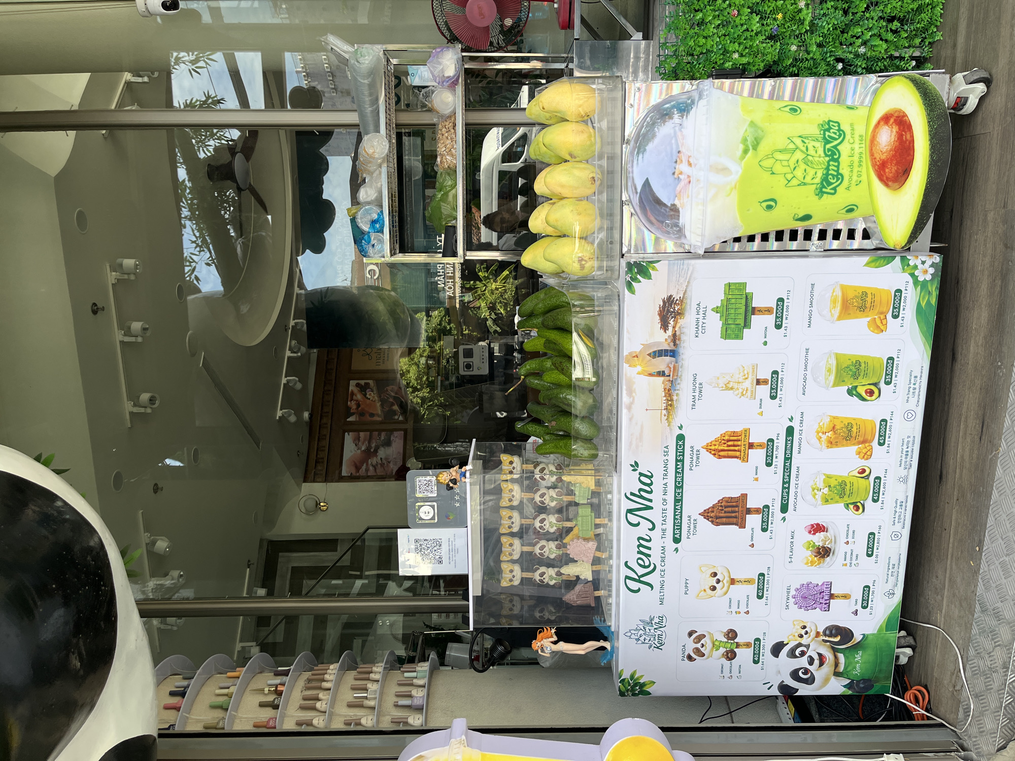 This is a street-level photograph of an outdoor ice cream stand called "Kem Nha" located in Nha Trang, Vietnam. The stand is positioned in front of a shop with a large glass window displaying various items for sale, including bottles of nail polish and other products that are not clearly identifiable. The ice cream stand itself is decorated with colorful signs and images of ice cream products.
Prominently displayed on the stand is a menu featuring different types of ice cream sticks and drinks, such as "Panda," "Puppy," "Ponagar Tower," "Tram Huong Tower," "Skywheel," and various smoothies. The prices are listed in Vietnamese Dong, US Dollars, Korean Won, and Philippine Peso. There are also visual representations of some of the offerings, including a large avocado next to a smoothie.
In front of the stand, there are several ice cream popsicles shaped like cartoon characters and local landmarks. Fresh produce, specifically avocados and yellow fruits resembling mangoes or papayas, are displayed in clear plastic containers. Behind the stand, some shelves hold various packaged goods and supplies. A red electric fan is visible to the right, and a security camera is mounted on the building. The overall scene suggests a commercial area, likely a busy street in Nha Trang, with an emphasis on promoting local treats and beverages.