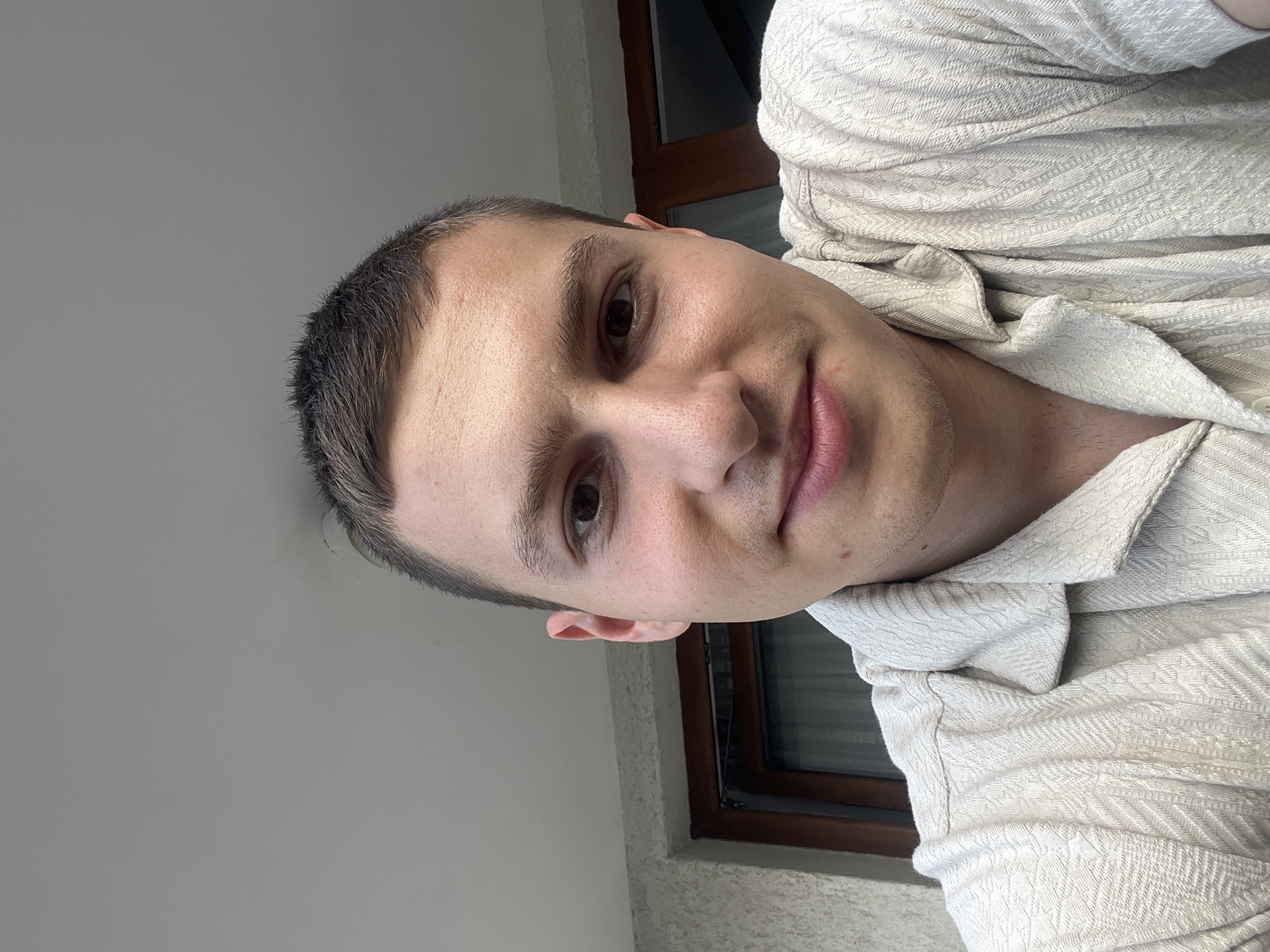 A close-up, medium-angle selfie shot of a young man. The man has dark hair, short-cropped, and light skin. He is wearing a light-colored shirt with a subtle textured pattern. The background shows a window with a brown frame, a portion of a gray wall, and potentially part of a building exterior. There is no evidence to definitively suggest that the image was taken in Varna, Bulgaria. Further details such as landmarks or specific architectural styles would be needed to confirm the location.
