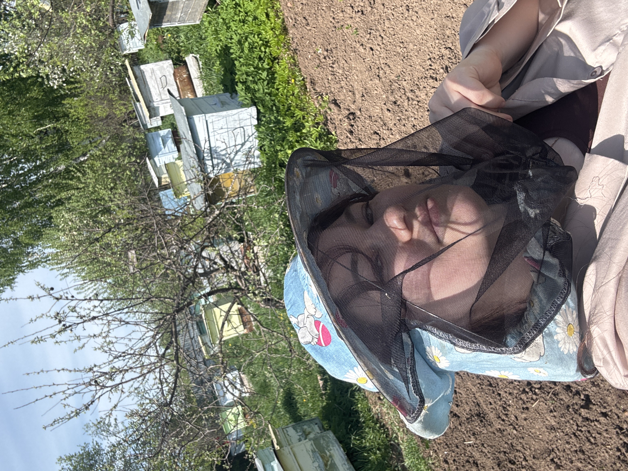 A person wearing a beekeeping veil with a light blue hat underneath, adorned with cartoon figures and daisies, stands in a garden setting. The background reveals a collection of white beehives dispersed among greenery, a bare tree with visible branches, and cultivated soil. The location is Маркидоновка, Russia.