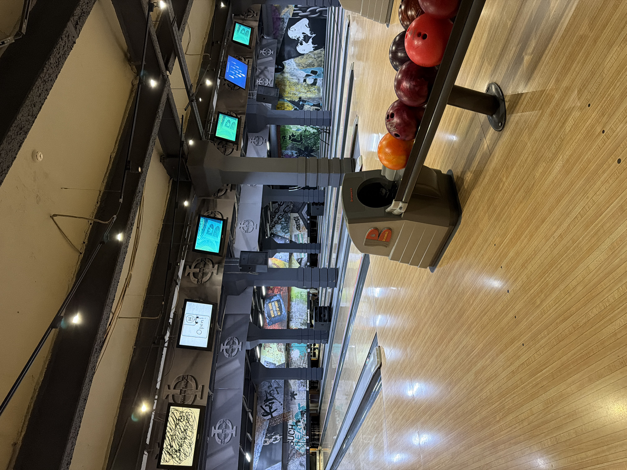 The image shows an interior view of a bowling alley with multiple lanes. The lanes are made of polished wood, with dark lines indicating the boundaries and foul lines. On the right side of the image, a bowling ball return rack is visible, loaded with several bowling balls of various colors, including orange, red, and dark purple. The balls are resting on a black angled surface. In the background, large pillars support an overhead structure. Above the lanes, several screens are mounted on the ceiling structure, displaying different graphics and text, including one screen with the word "Open!". The walls behind the lanes are decorated with graffiti-style murals. The lighting is functional, with spotlights illuminating the bowling lanes. There are no people visible in the immediate foreground or on the lanes. The overall impression is of a modern entertainment venue. The location is identified as Moscow, Russia.