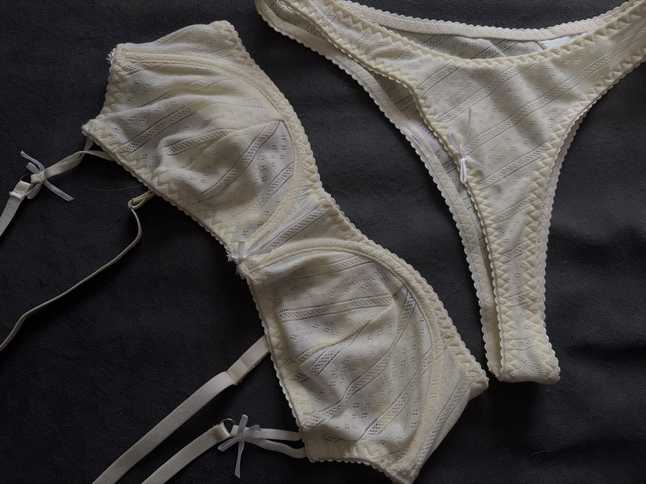 A matching lingerie set in a light, off-white color is arranged on a dark gray surface. The bra is a soft-cup style with delicate lace detailing and adjustable straps adorned with small bows. A matching thong with similar lace and a small bow is positioned next to the bra. The set appears lightweight and feminine, designed with comfort in mind.