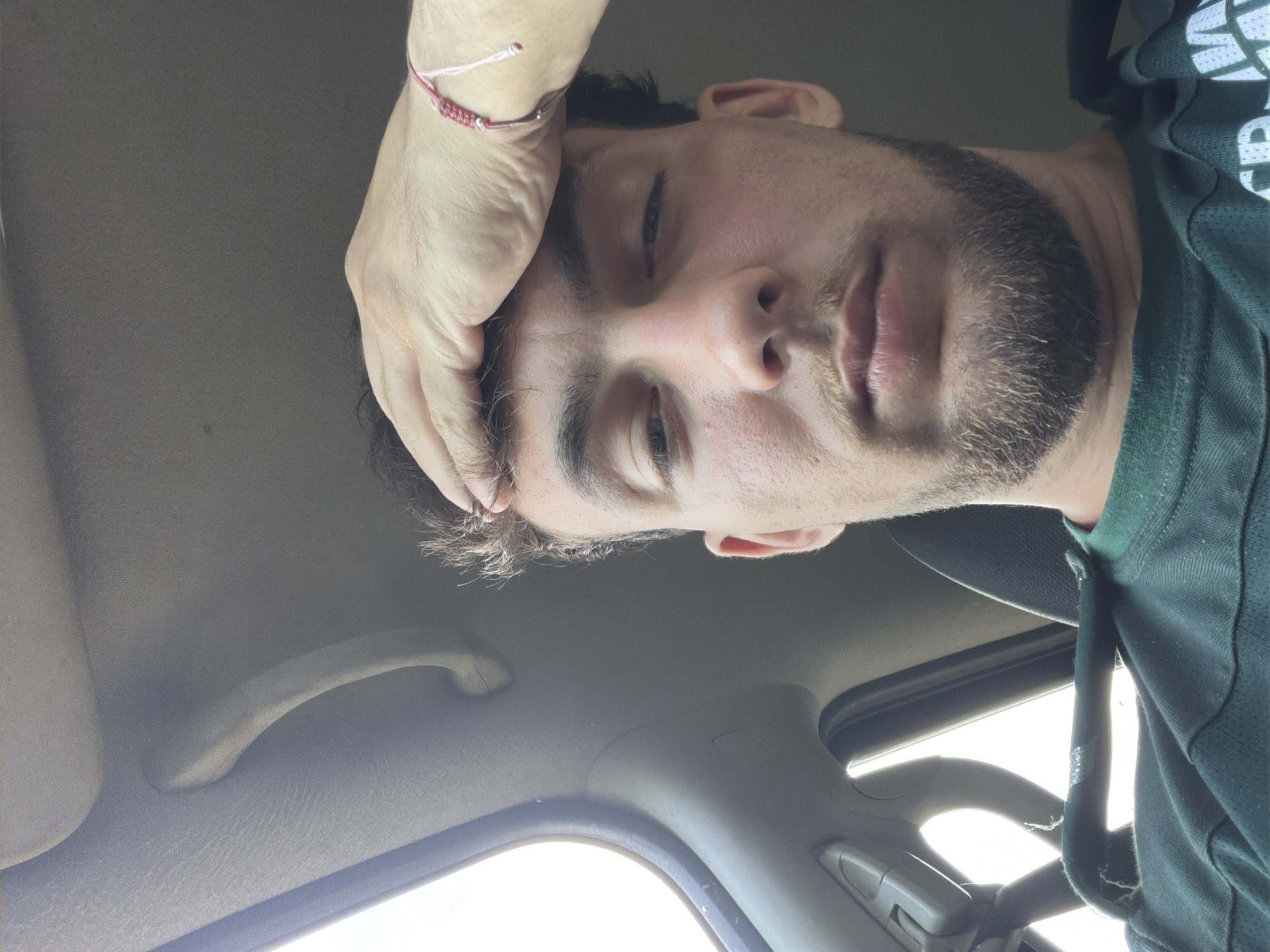 A male subject is seated inside a vehicle in Rezovo, Bulgaria. He has dark hair, a light beard and mustache, and looks directly at the camera with a heavy-lidded, neutral expression. His right hand is placed on his forehead, and a red and white braided bracelet is visible on his right wrist. He is wearing a dark green shirt. The vehicle's interior, likely a car, features a light-colored headliner and a passenger grab handle visible on the left side. Bright daylight is entering through the left window, creating an overexposed area.