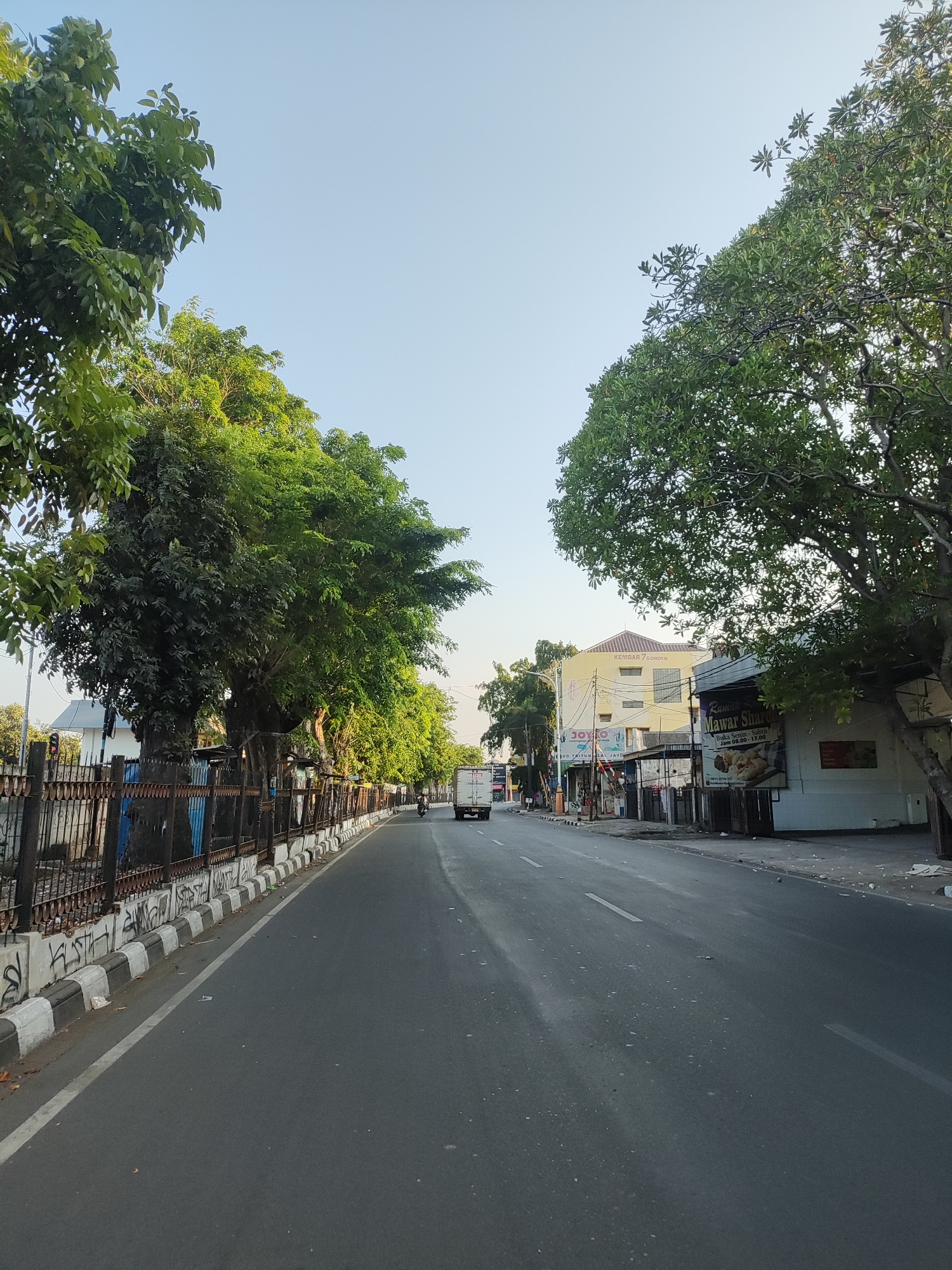 The image captures a wide asphalt road with white lane markings in Cirebon, Indonesia. The road is flanked by lush green trees on both sides, with one side featuring a metal fence and a sidewalk. Buildings, including a yellow structure with the text "KEMBAR / COROYN" and a white building with an advertisement for "Mawar Sh..." are visible along the street. A white truck and a motorcycle are present on the road. The sky is clear and blue, indicating a bright day. The overall scene depicts a typical urban streetscape in Indonesia.