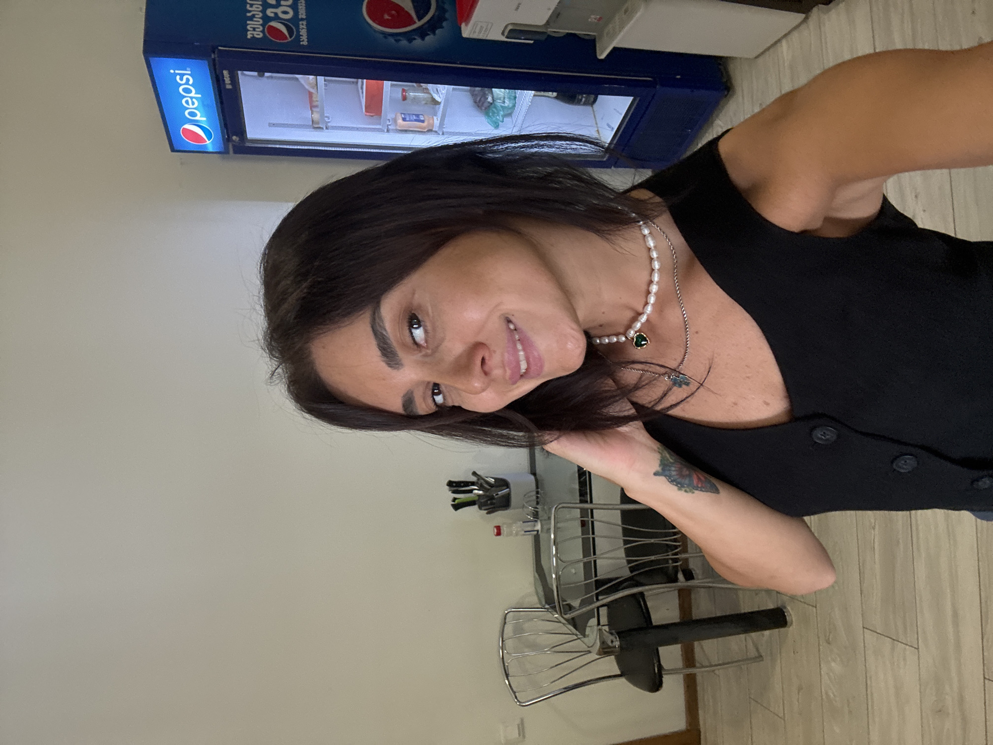 A dark-haired woman with a light complexion and brown eyes is captured in a selfie. She is wearing a black vest-like garment adorned with dark buttons and a pearl necklace with a green heart pendant. A tattoo is visible on her left forearm. The background reveals a commercial setting, likely a cafe or convenience store, in Tbilisi, Georgia. To her left, a glass-topped table with two chrome chairs is set up, with a container of utensils and a bottle on it. To her right, a blue Pepsi refrigerator is visible, displaying various drinks and products inside. A water dispenser is also present next to the refrigerator. The flooring is a light-colored wood-effect laminate. The overall lighting suggests an indoor environment.