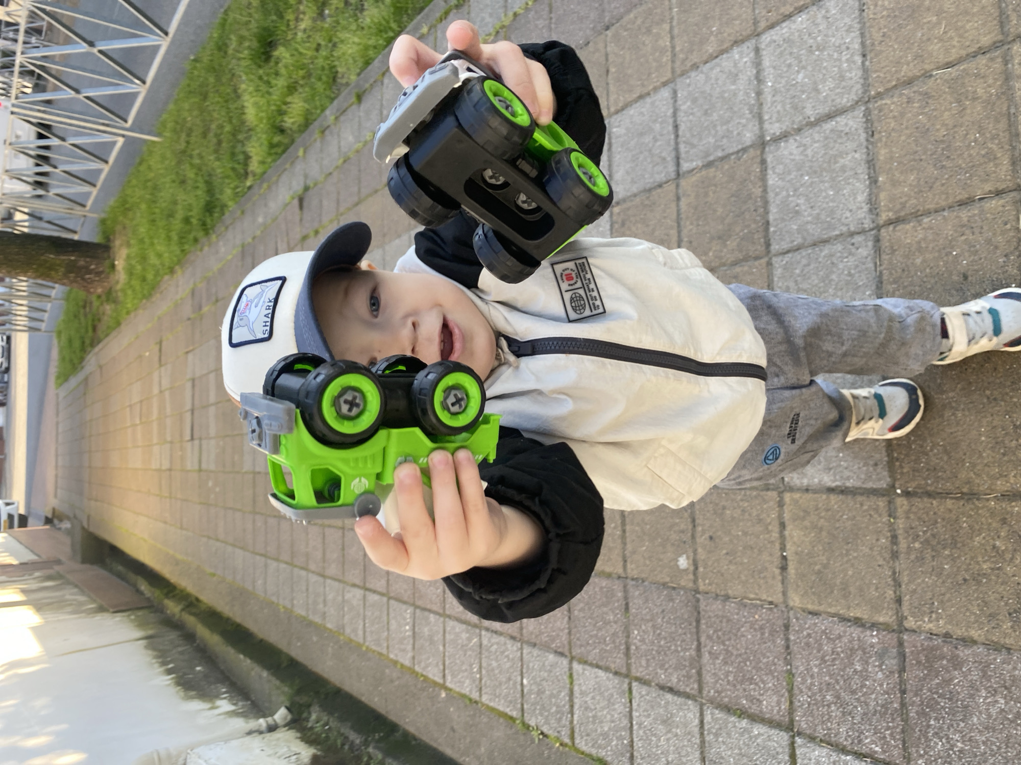 The image features a young child, likely a boy, playing outdoors in Sochi, Russia, on a sunny day. The child, appearing to be between three and six years old, looks directly at the camera with a wide, happy smile. He is holding up two identical or very similar toy monster trucks, one in each hand, with bright green wheels and chassis contrasting with black bodies.
The child wears a white baseball cap with a "SHARK" patch featuring a shark graphic, a white and black zip-up jacket with a small patch displaying a globe-like graphic and some indistinct text, and grey sweatpants with a visible logo.
The background shows a paved area, likely a sidewalk or pathway, composed of square concrete tiles in varying shades of grey and brown. Alongside the pathway, there's a strip of green grass and a tree trunk visible. Further back, light-colored concrete or brick walls and some metal structures can be seen, suggesting an urban or park setting. The bright natural light indicates it's daytime, and the child's expression conveys joy and playfulness.