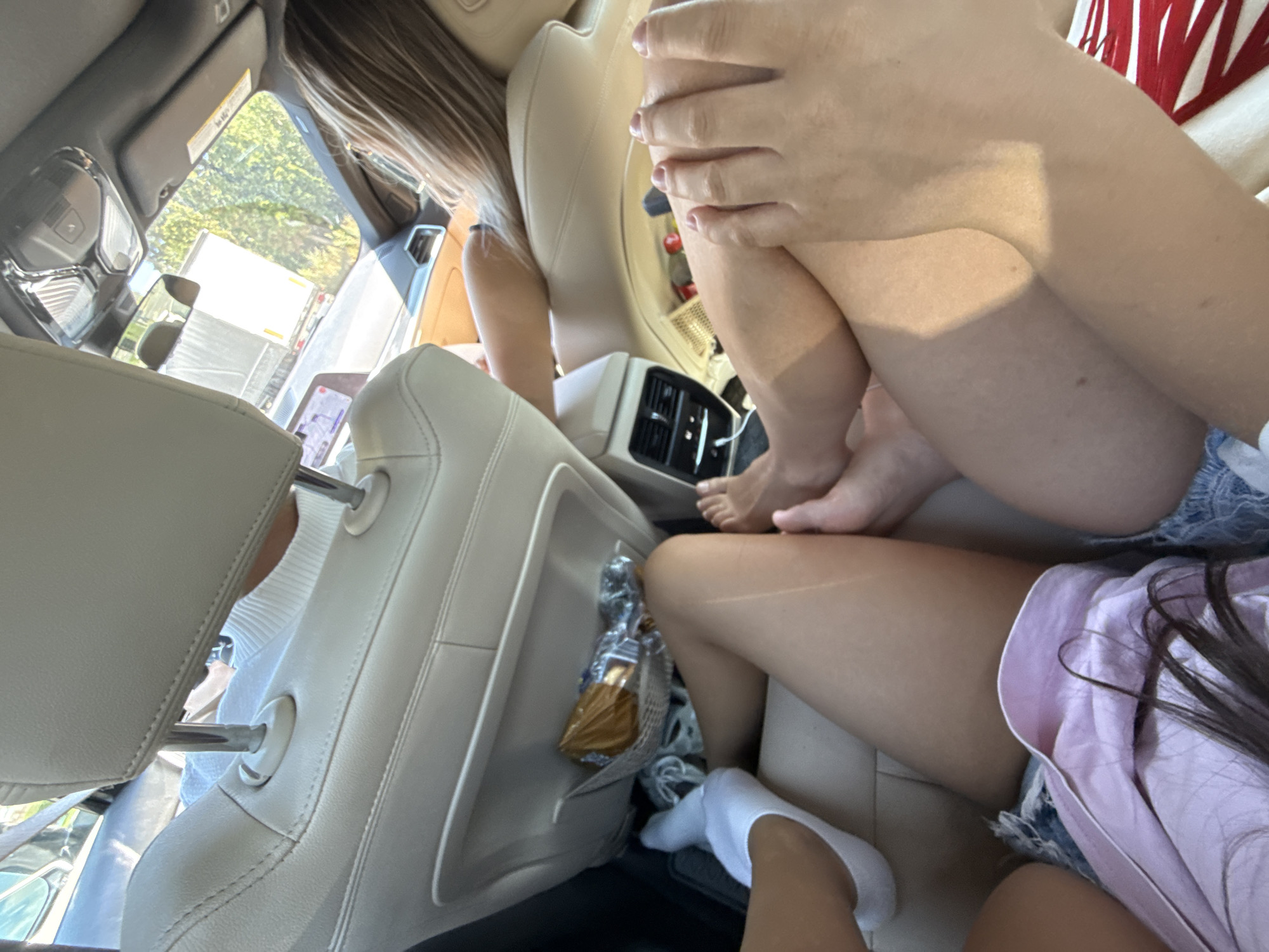 The media file captures a candid moment inside a vehicle, likely taken on a sunny day, as indicated by the bright natural light. The scene is set within the interior of a car, focusing on the legs and arms of at least two individuals. The interior appears to be upholstered in light-colored leather, with visible headrests and seats. In the background, through the car window, glimpses of greenery and trees suggest an outdoor setting, possibly a road trip.
One person's legs are visible, with one foot wearing a white sock, resting on the floor of the car. Another person's thighs are prominently featured, crossed at the knee, with one hand resting on their leg. The individual's skin shows a tan line, suggesting sun exposure. A third person's arms and hands are also visible, with manicured fingernails. They are wearing what appears to be denim shorts. A pink garment is also visible, possibly a shirt.
Tucked into a seat pocket is a wrapped item, possibly a snack, encased in a shiny, golden wrapper and a mesh bag. The overall impression is one of casual comfort and relaxation during travel. There is no visible text in the image, and the emotions conveyed are relaxed and informal. The setting suggests a warm weather outing, possibly a summer journey.