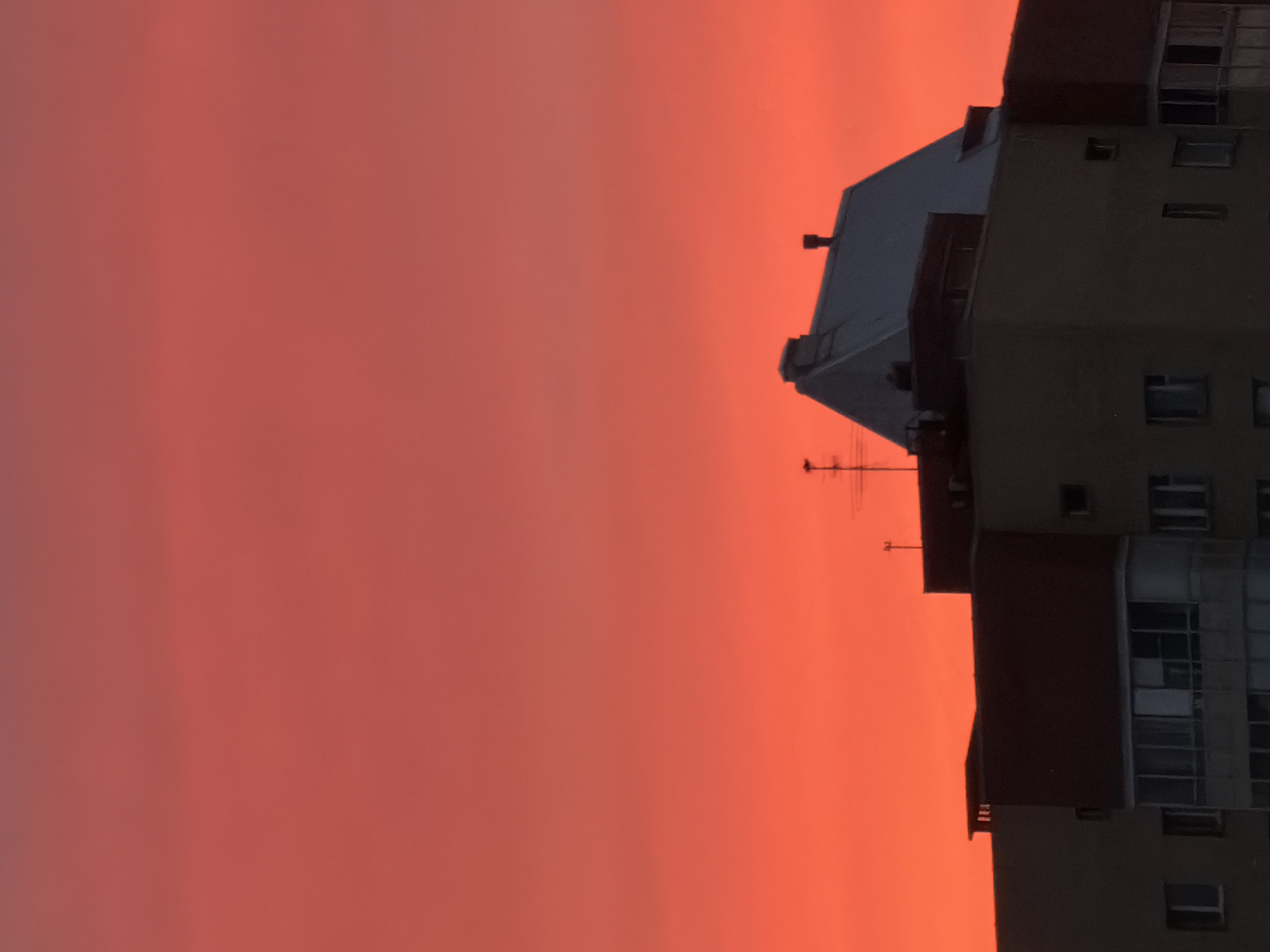 The image shows a tall building against a vivid, reddish-orange sunset. The building has multiple stories with numerous windows. An antenna is visible on the roof. The location is likely in a city, based on the presence of the building, and its association with the city of Saint Petersburg, Russia.