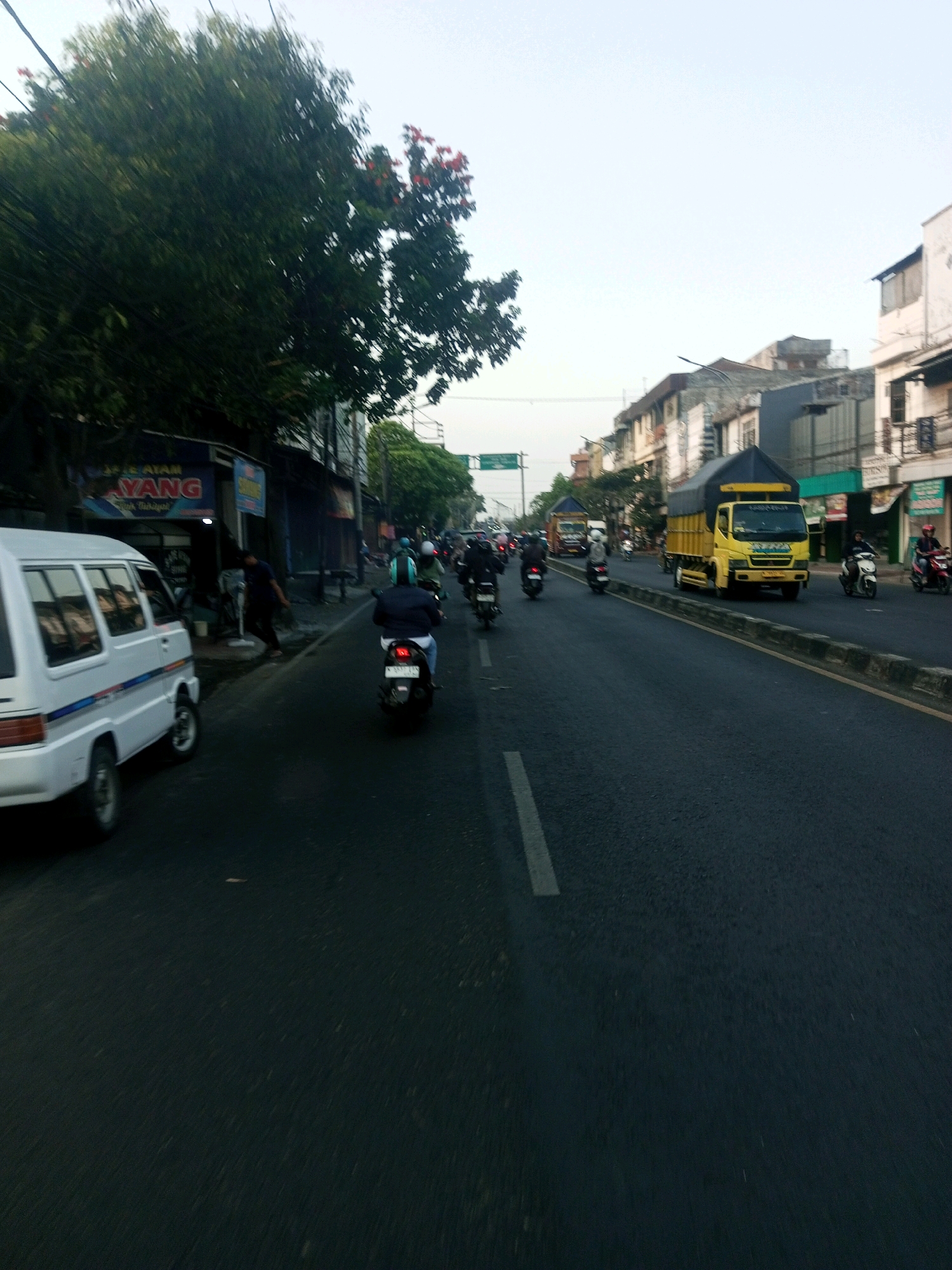 The image captures a street scene in the city of Malang, Indonesia, likely during daytime. A white van is parked on the left side of the road. Several motorbikes are in motion on the asphalt road, with riders wearing helmets. One rider in particular is visible in the foreground, on a black motorbike, wearing a dark jacket and blue jeans. Further down the road, a yellow truck with a dark tarp covering its cargo is prominently visible. Buildings line the right side of the street, displaying a mix of architectural styles. Trees with green foliage are also present, adding to the urban landscape. The sky above is overcast.