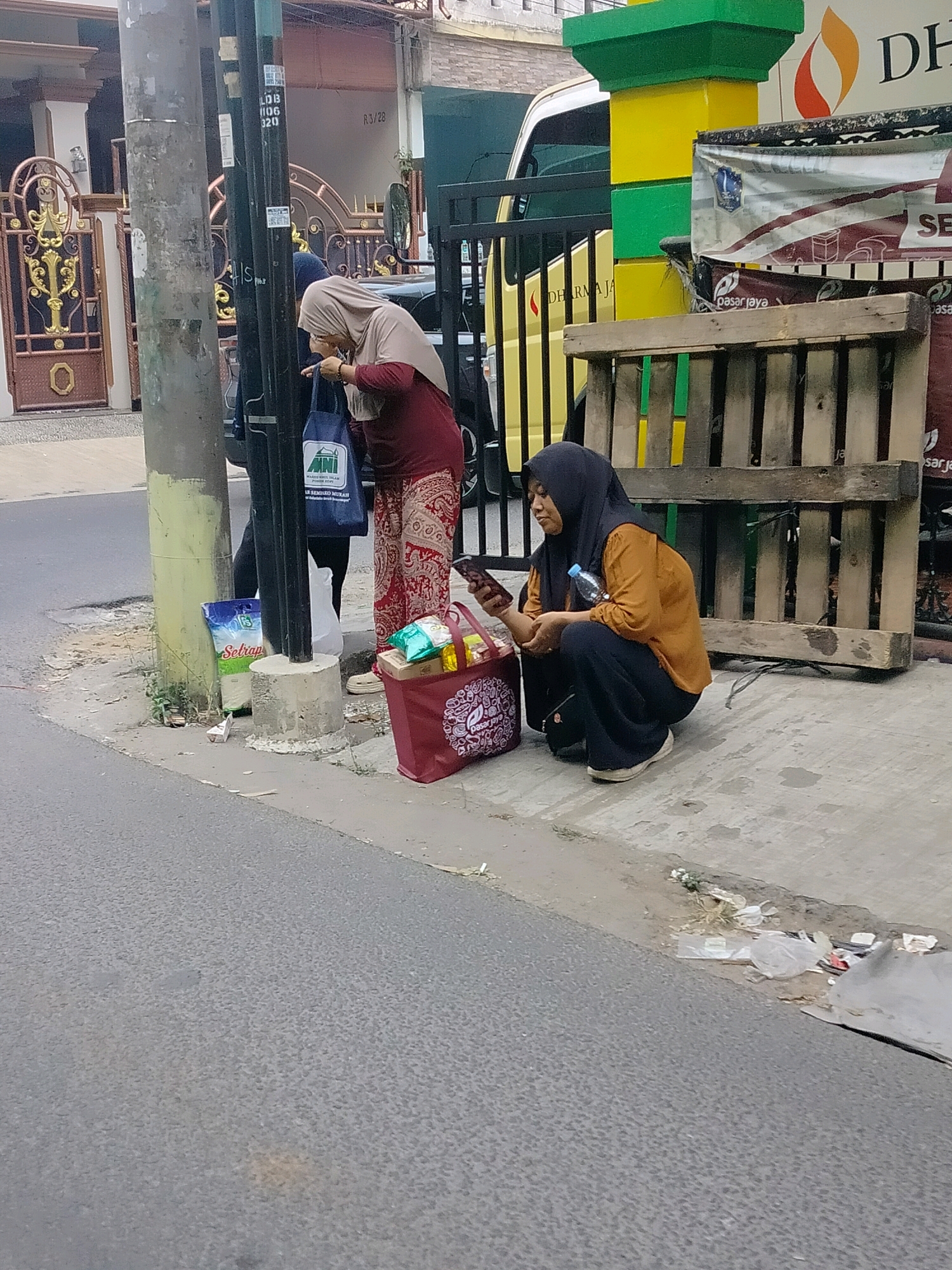 In East Jakarta, Indonesia, two veiled women are depicted near a street pole and a wooden pallet structure. The woman on the left, wearing a beige hijab and a maroon top with a patterned skirt, is pointing at something on her phone, which she holds in her right hand. She stands next to a blue tote bag. To her right, the second woman, with a dark hijab and an orange top over dark pants, is crouched down, looking at her smartphone. A plastic water bottle is visible near her. In front of her is a red tote bag filled with grocery items. A bag of "Setrap" detergent is placed on the ground near the street pole. The scene is set on a concrete sidewalk adjacent to a road, with a building and vehicles visible in the background. Debris and litter are scattered on the ground.